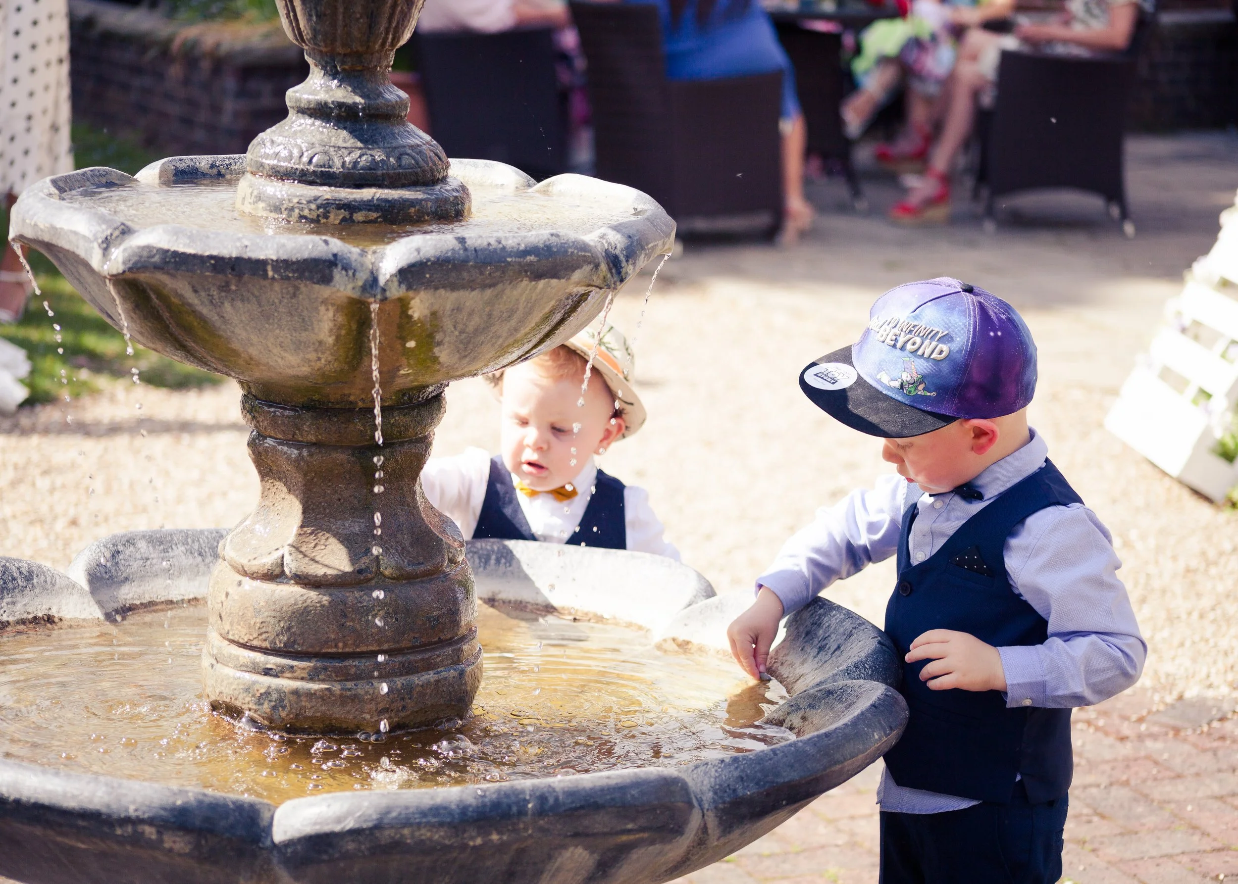 Two children, a boy and a girl, playing in a water fountain outdoors during daytime. The boy is wearing a blue hat with a graphic and a light blue shirt, and the girl is wearing a navy dress with white accents. They are reaching into the water at the
