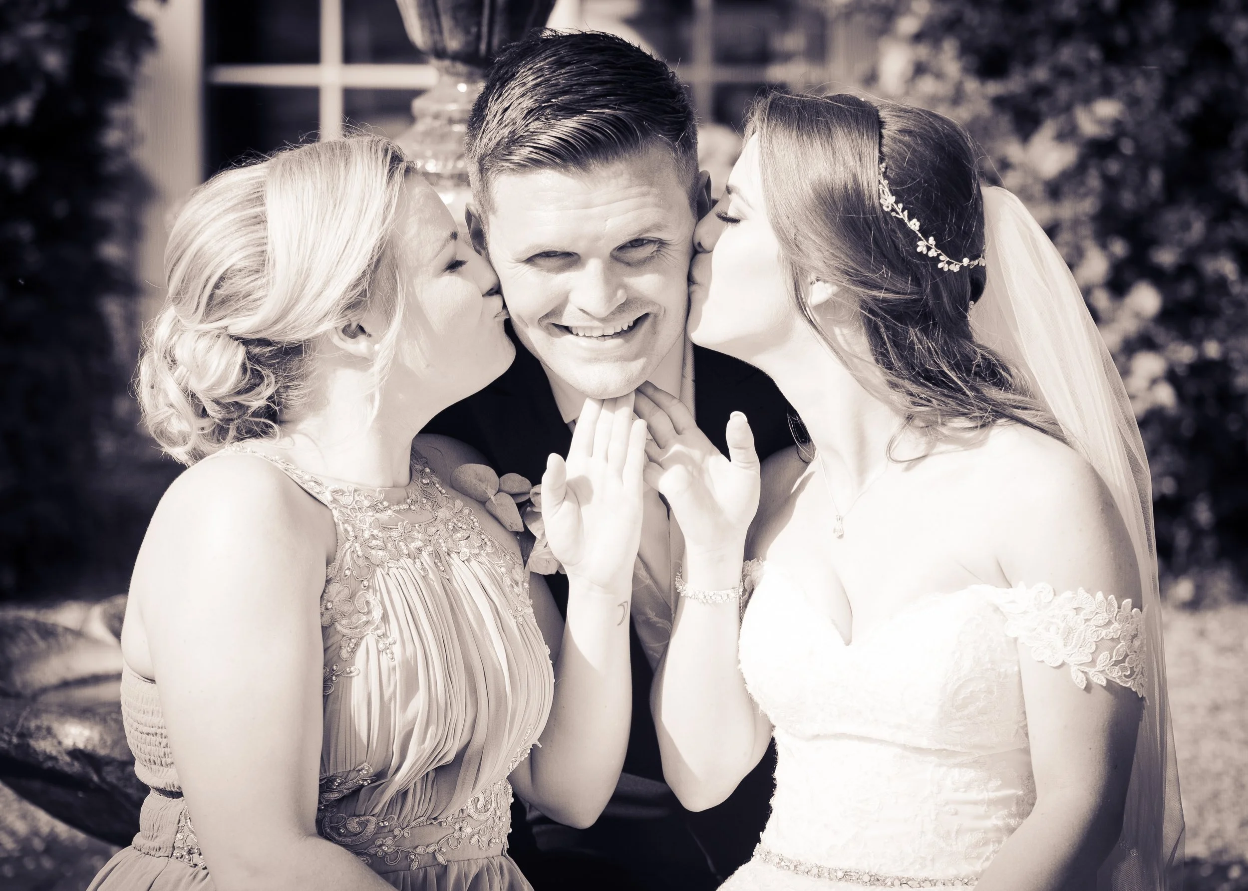 A groom is flanked by two brides, each giving him a kiss on the cheek, during a wedding celebration outside. The photo is in black and white.