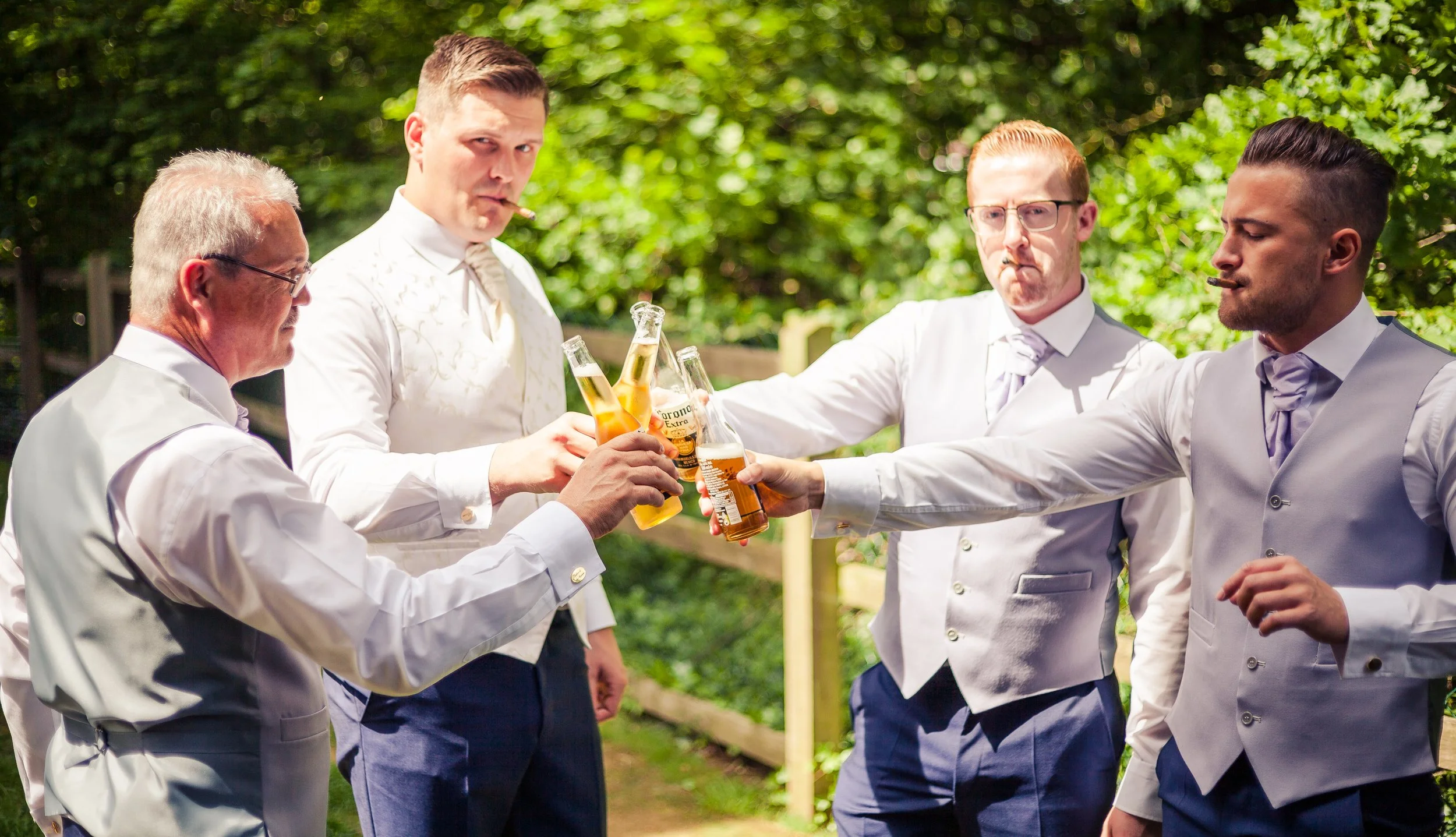 Group of men in white suits clinking beer bottles at outdoor gathering.