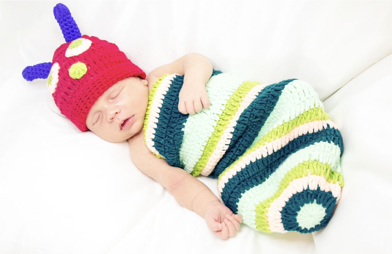 A sleeping baby wearing a colorful crocheted hat with antennae and a striped crocheted blanket, lying on a white surface.