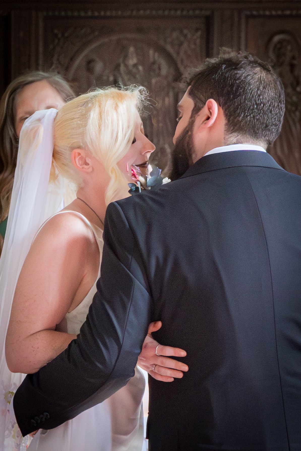 A bride and groom standing close, gazing at each other during their wedding ceremony, with a woman partially visible in the background and ornate wooden carvings in a church or cathedral.