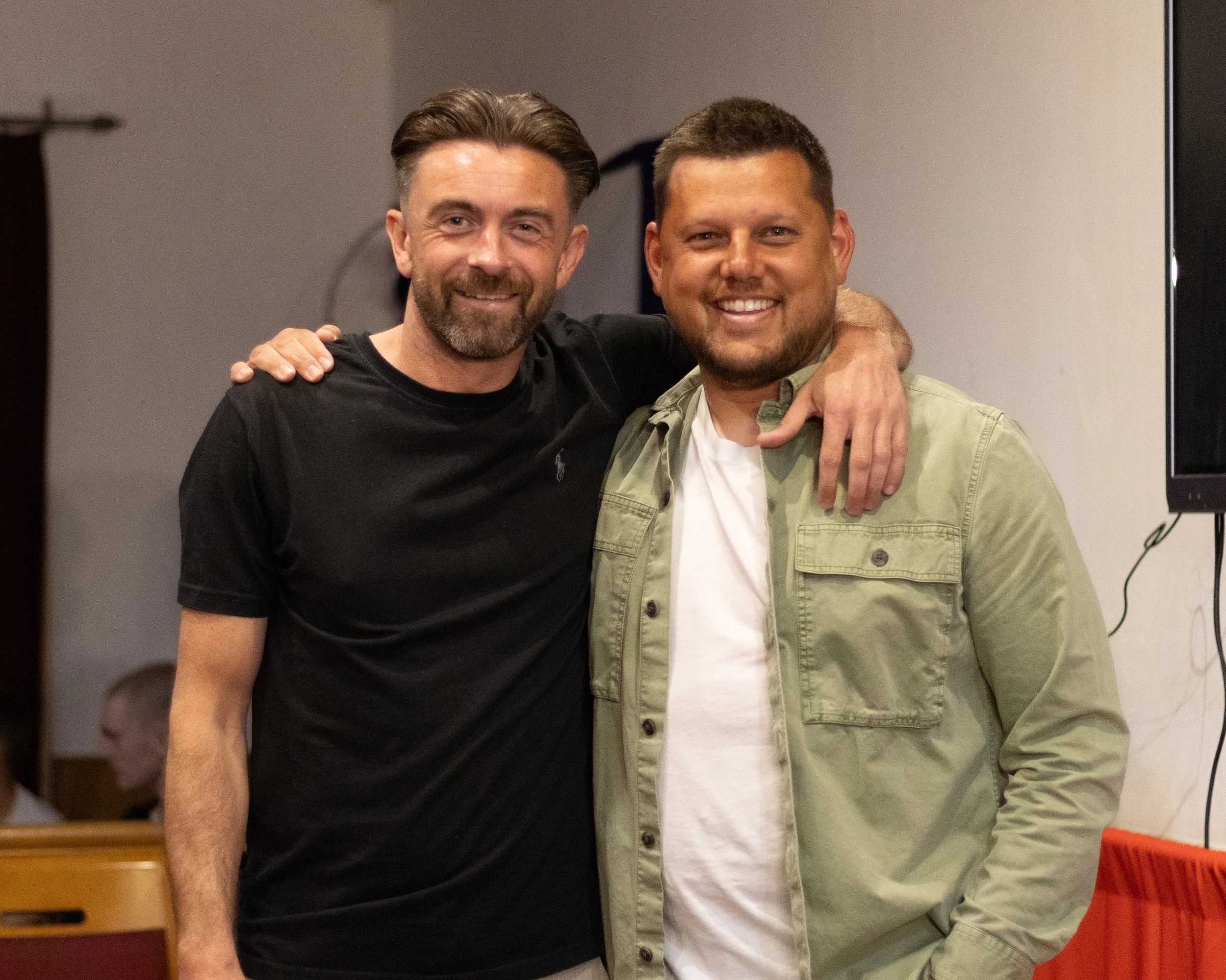 Two men smiling and embracing each other indoors, with a plain wall and a large TV or monitor behind them.
