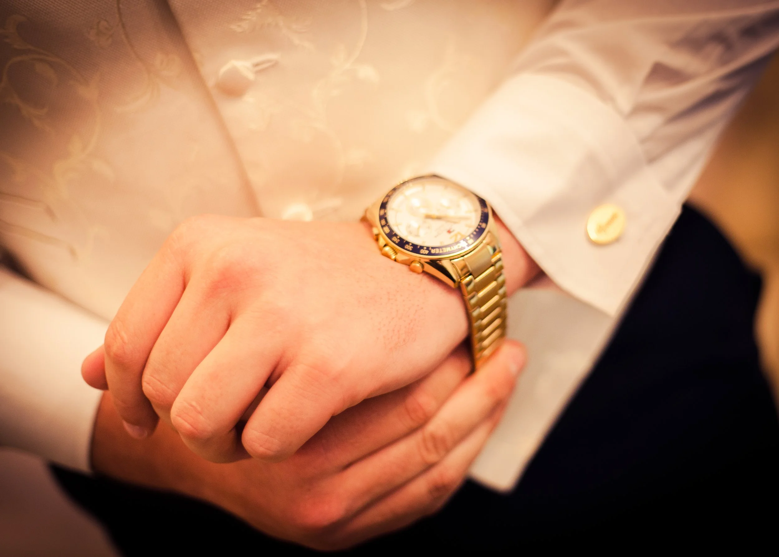 Person wearing a gold and silver wristwatch, dressed in a white shirt with a gold button, with their hand resting on their knee.