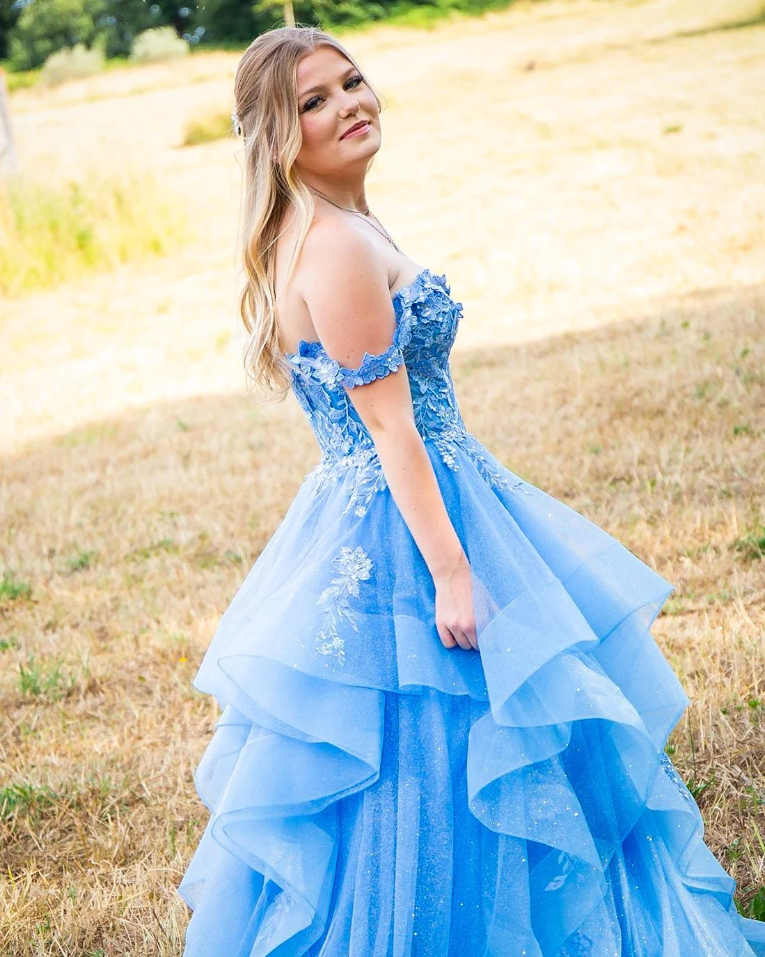 Young woman in a blue off-the-shoulder gown with floral embroidery, standing in a field with trees in the background.