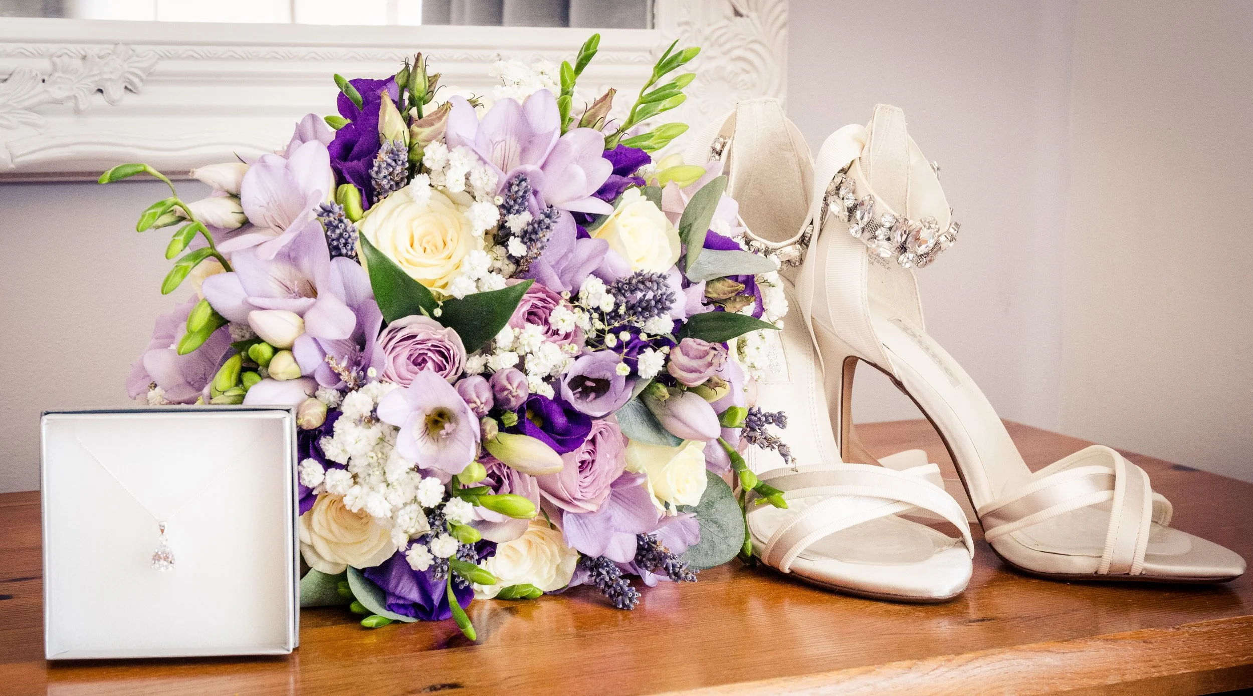 A large arrangement of purple, white, and cream flowers on a wooden surface with white high-heeled shoes decorated with large rhinestones, a white clutch purse, and a white ornate frame in the background.