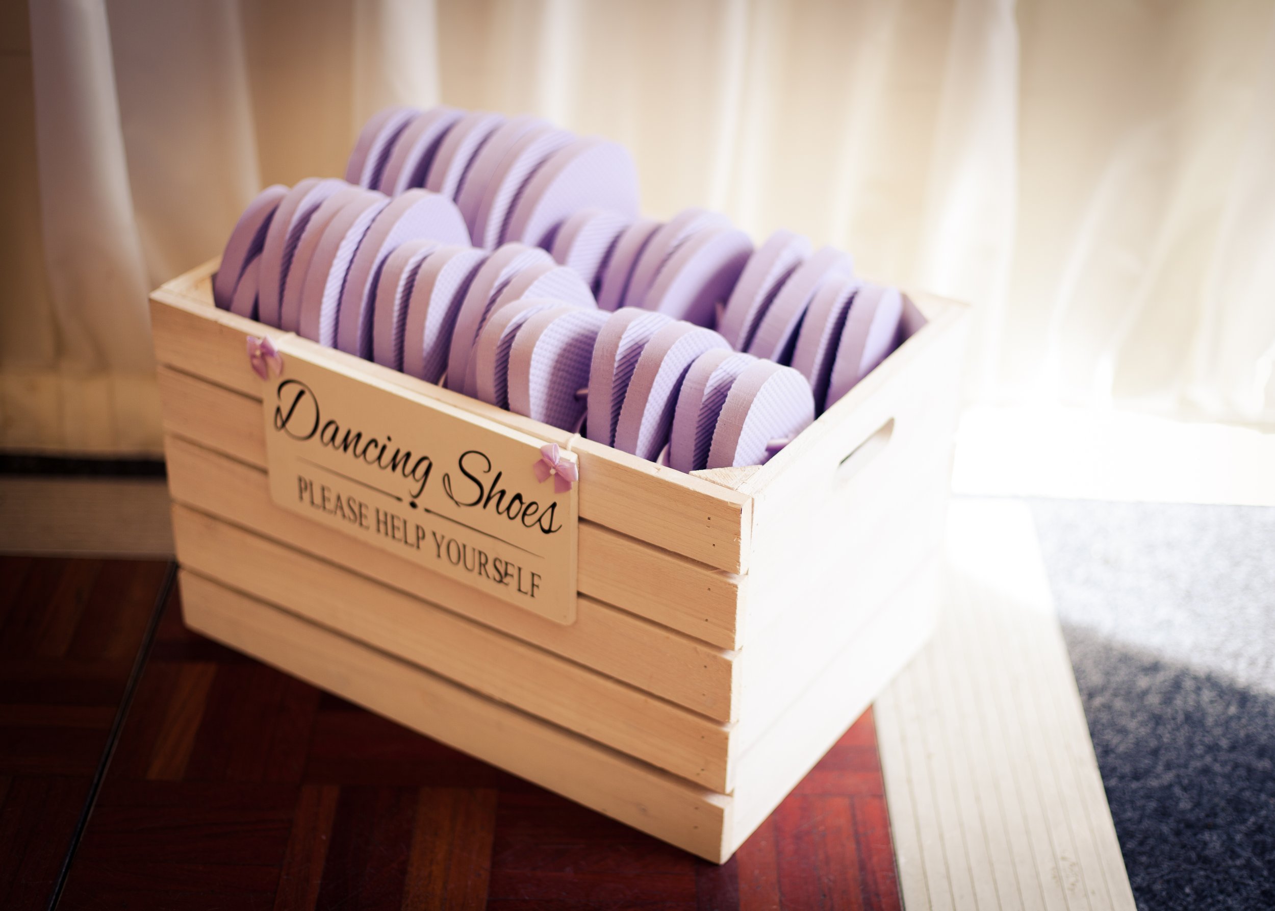 A wooden box labeled 'Dancing Shoes' contains rows of purple and white striped shoes, likely for a dance or wedding event, placed against a background of soft, light-colored curtains.