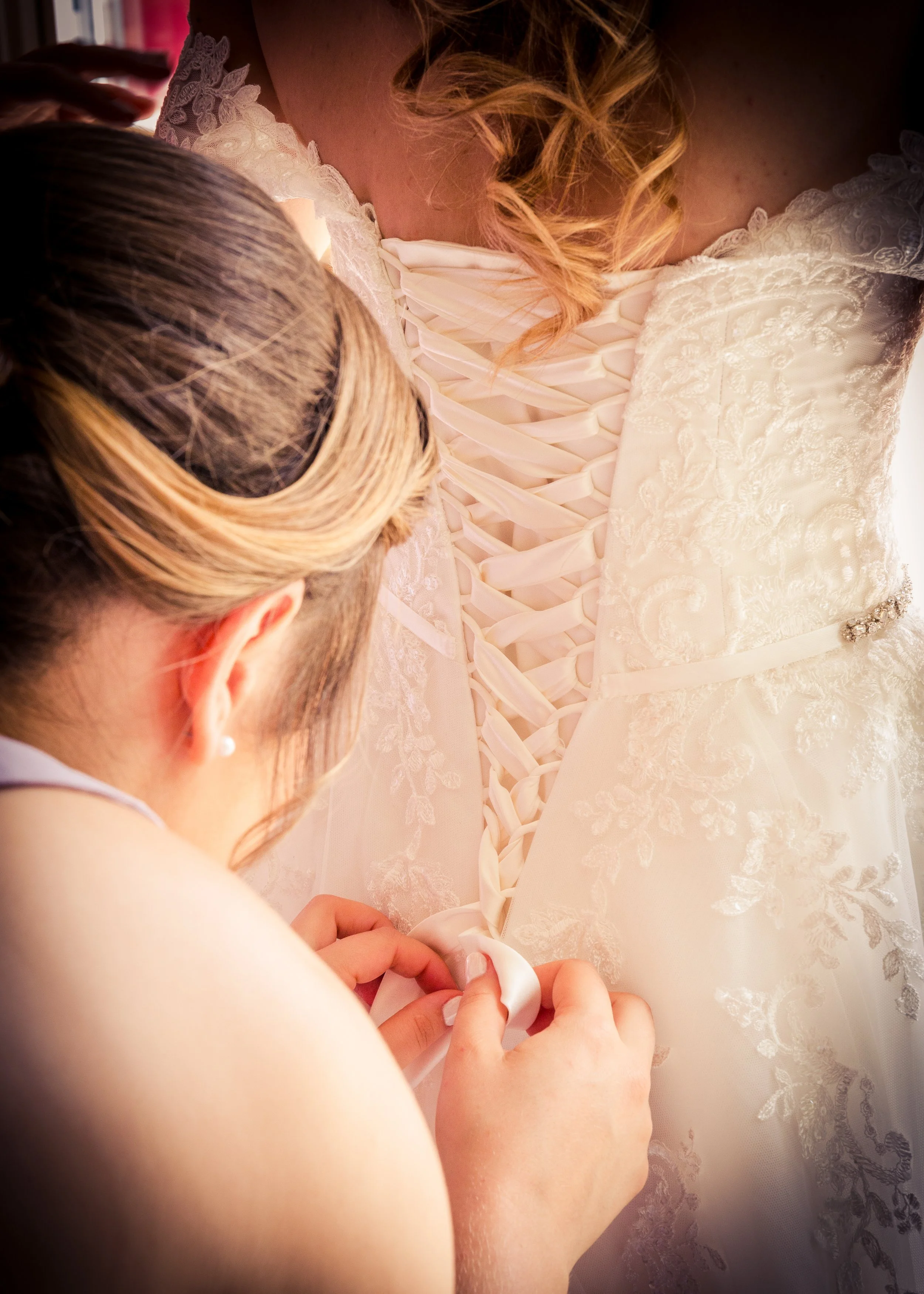 A person with light-colored hair in an updo is helping a bride lace up her wedding dress from the back. The wedding dress is white with intricate lace details.