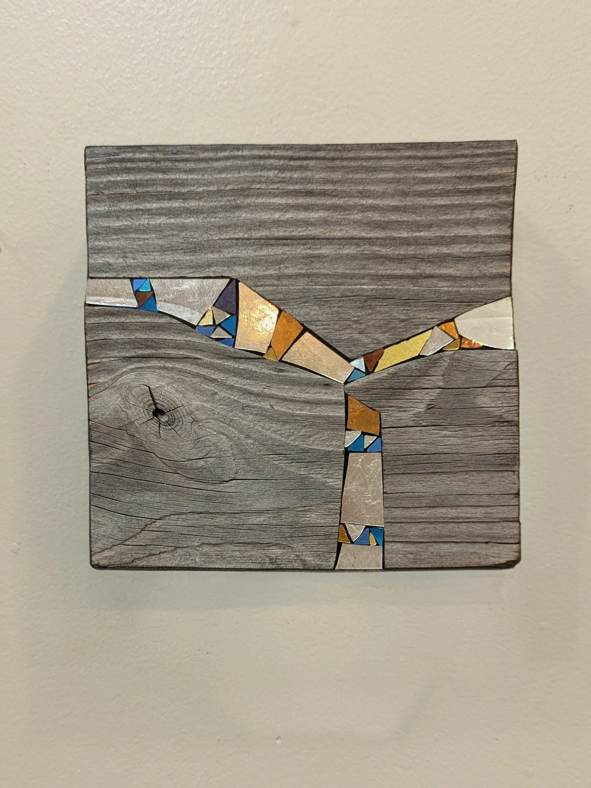 Une œuvre d'art composée de morceaux de bois gris, avec des fragments colorés en or, bleu, violet et marron intégrés dans une forme de serpent ou de coude dans le bois, fixé sur un fond blanc.