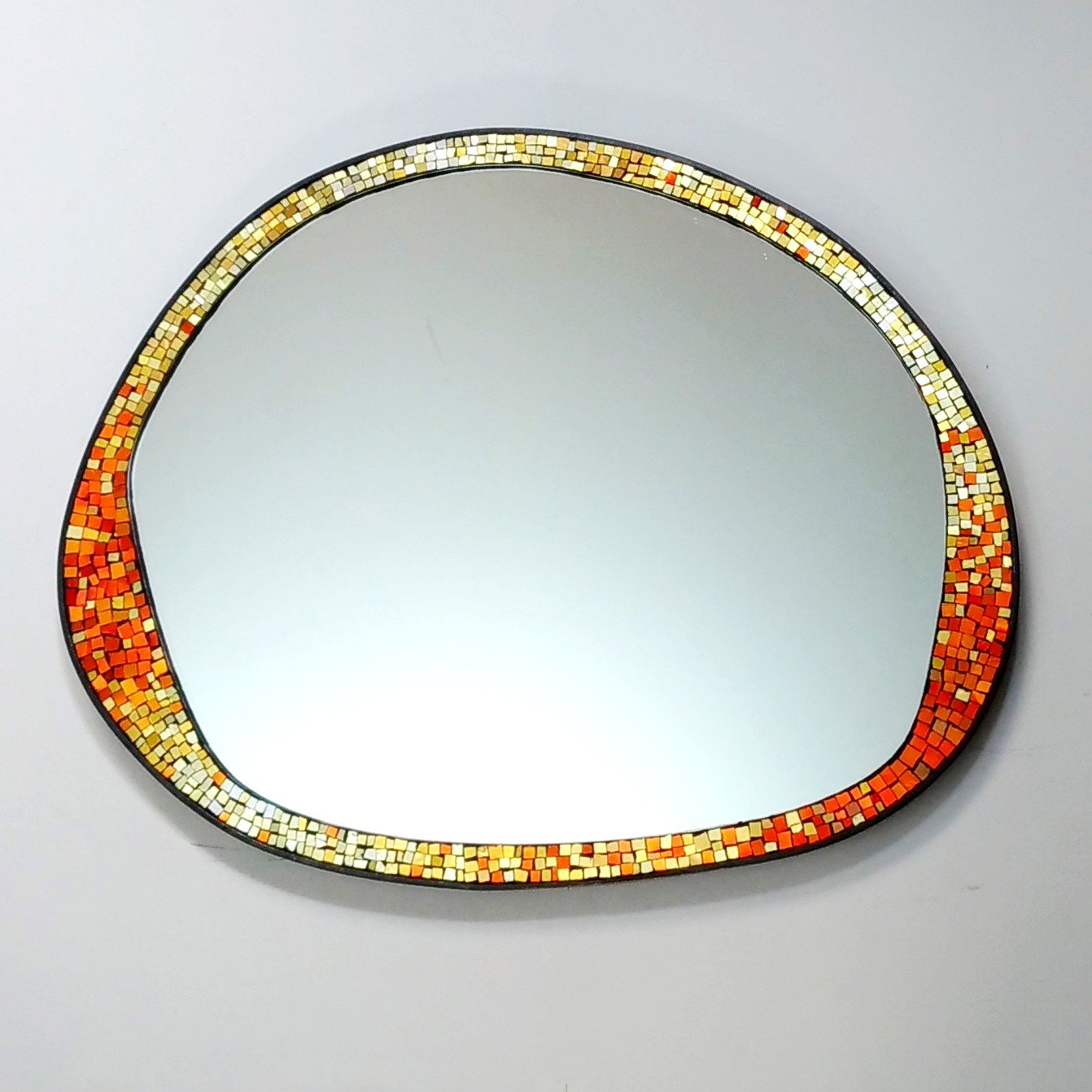 Un miroir ovale avec un cadre décoré de petits mosaïques aux couleurs chaudes, comme le rouge, l'orange, le jaune et le beige, posé sur une surface blanche.