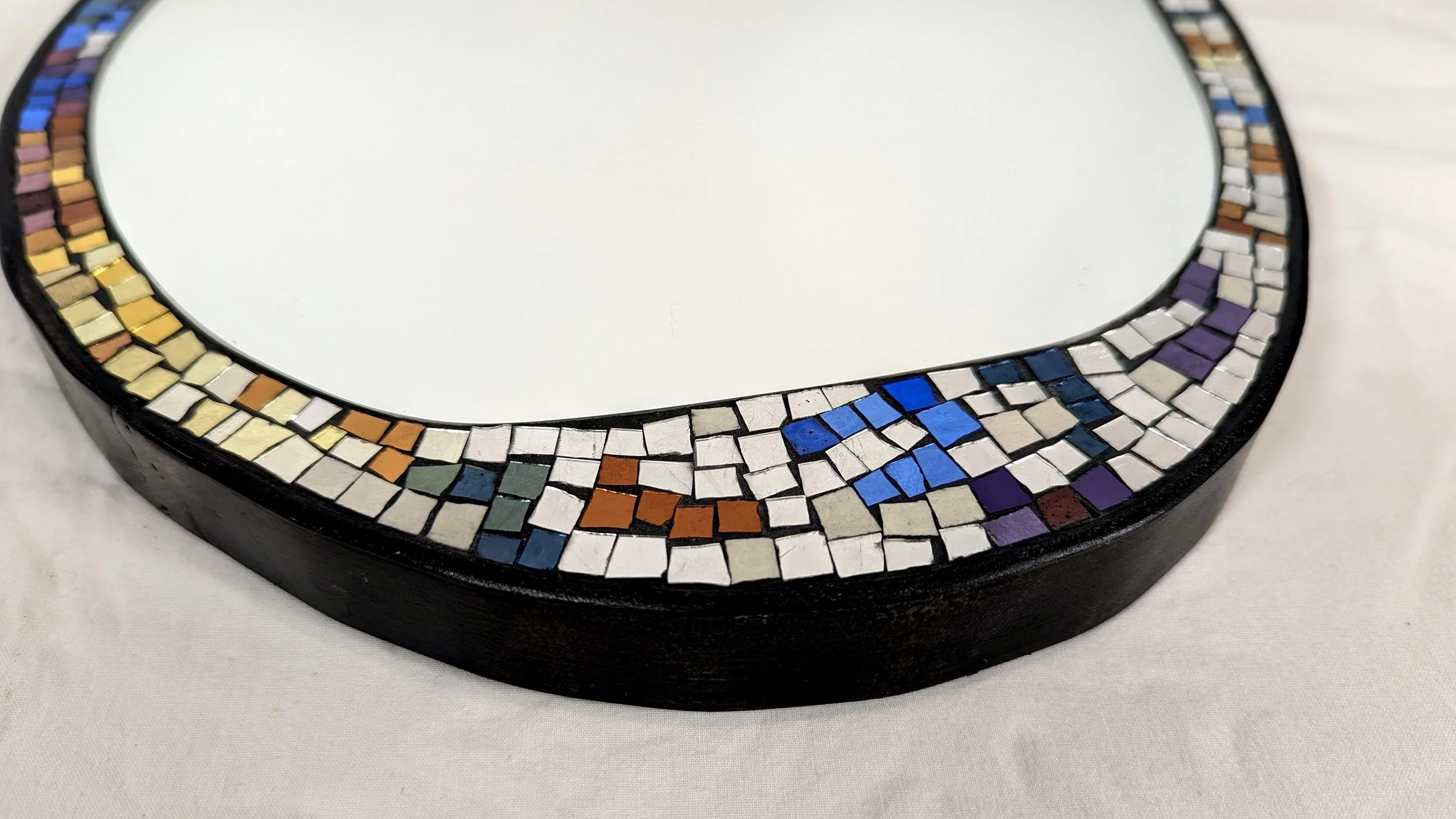 Miroir rond décoré avec une mosaïque de petits carrés colorés en shades de blanc, bleu, violet, jaune, beige, orange et gris, sur un fond blanc.