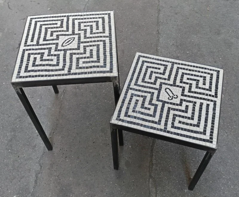 Deux tables avec des mosaïques en noir et blanc sur le dessus, chacune avec un labyrinthe et une forme de feuille au centre, placées sur un sol en béton.