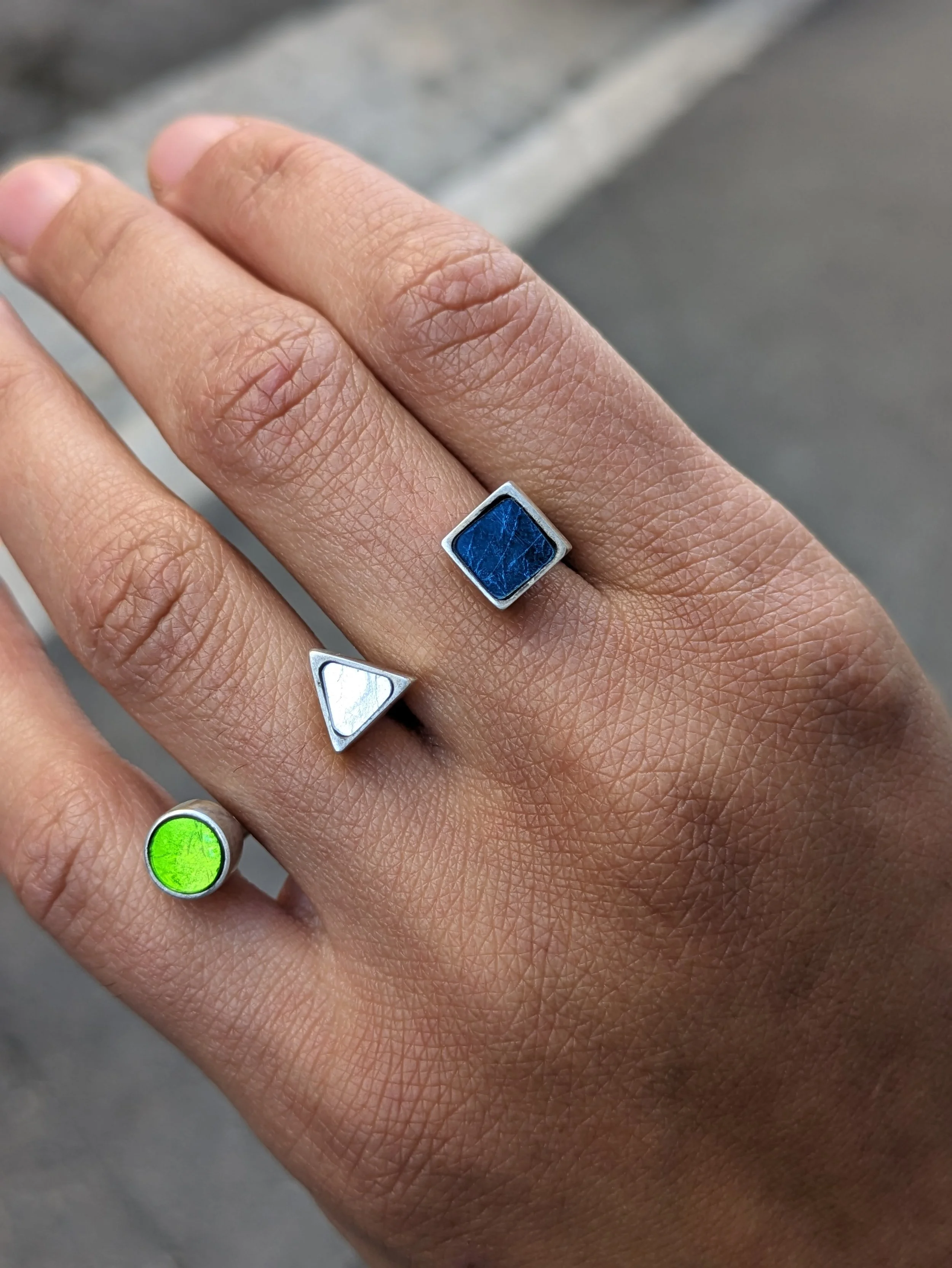 Image d'une main portant une bague double en mosaïque avec des formes géométriques distinctes, une bleu, une argentée en forme de triangle, et une verte
