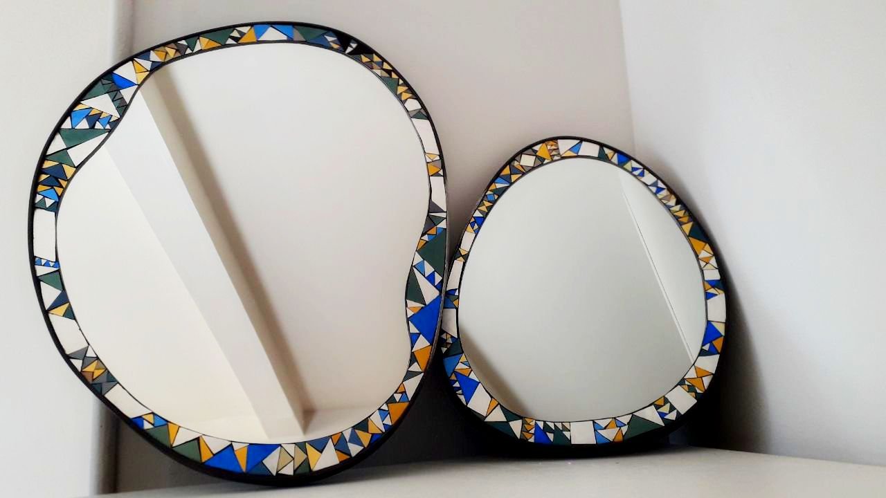 Deux miroirs ovales en mosaïque avec un cadre décoré de motifs colorés, posés contre un mur blanc.