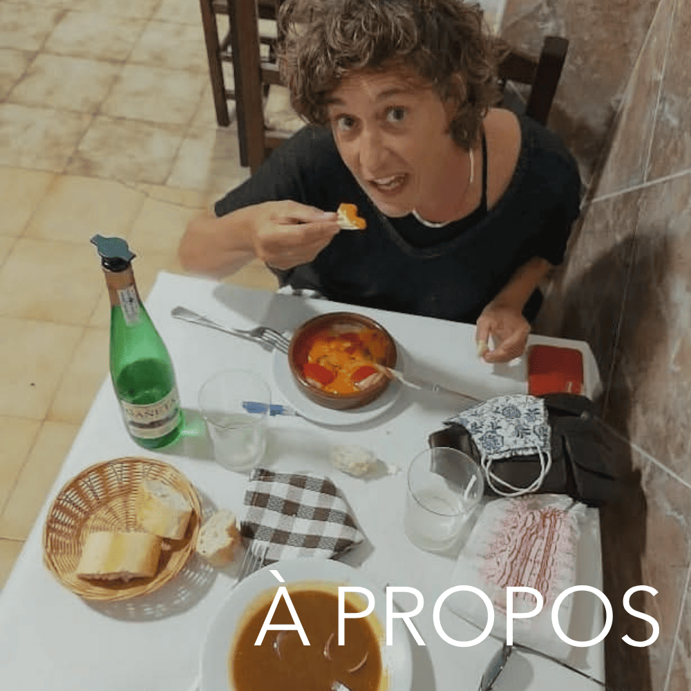 Une femme avec des cheveux bouclés courts mangeant une soupe dans un restaurant, assise à une table avec une bouteille d'eau gazeuse, des verres, du pain, et d'autres plats. La femme porte une chemise noire.