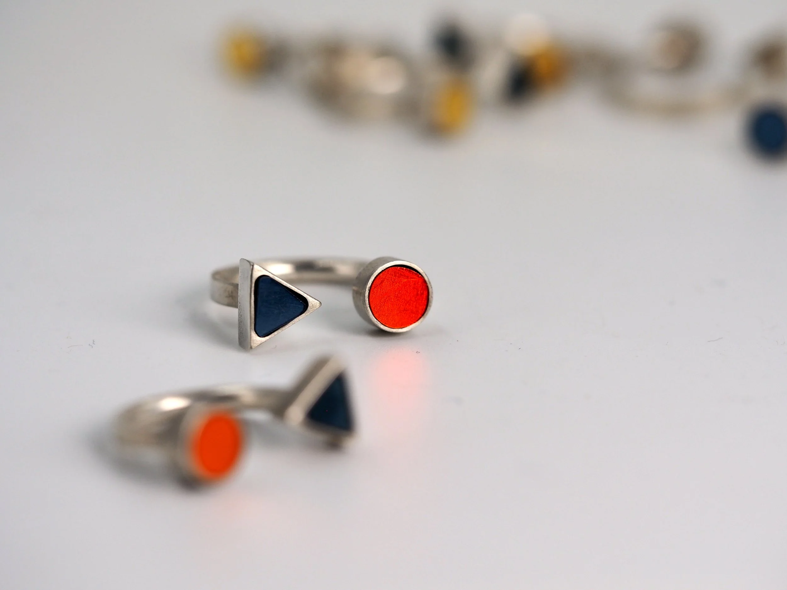 Bague en métal en mosaïque avec un design géométrique, comportant une partie triangulaire bleue et une partie circulaire orange, posée sur une surface blanche avec d'autres bijoux flous en arrière-plan.