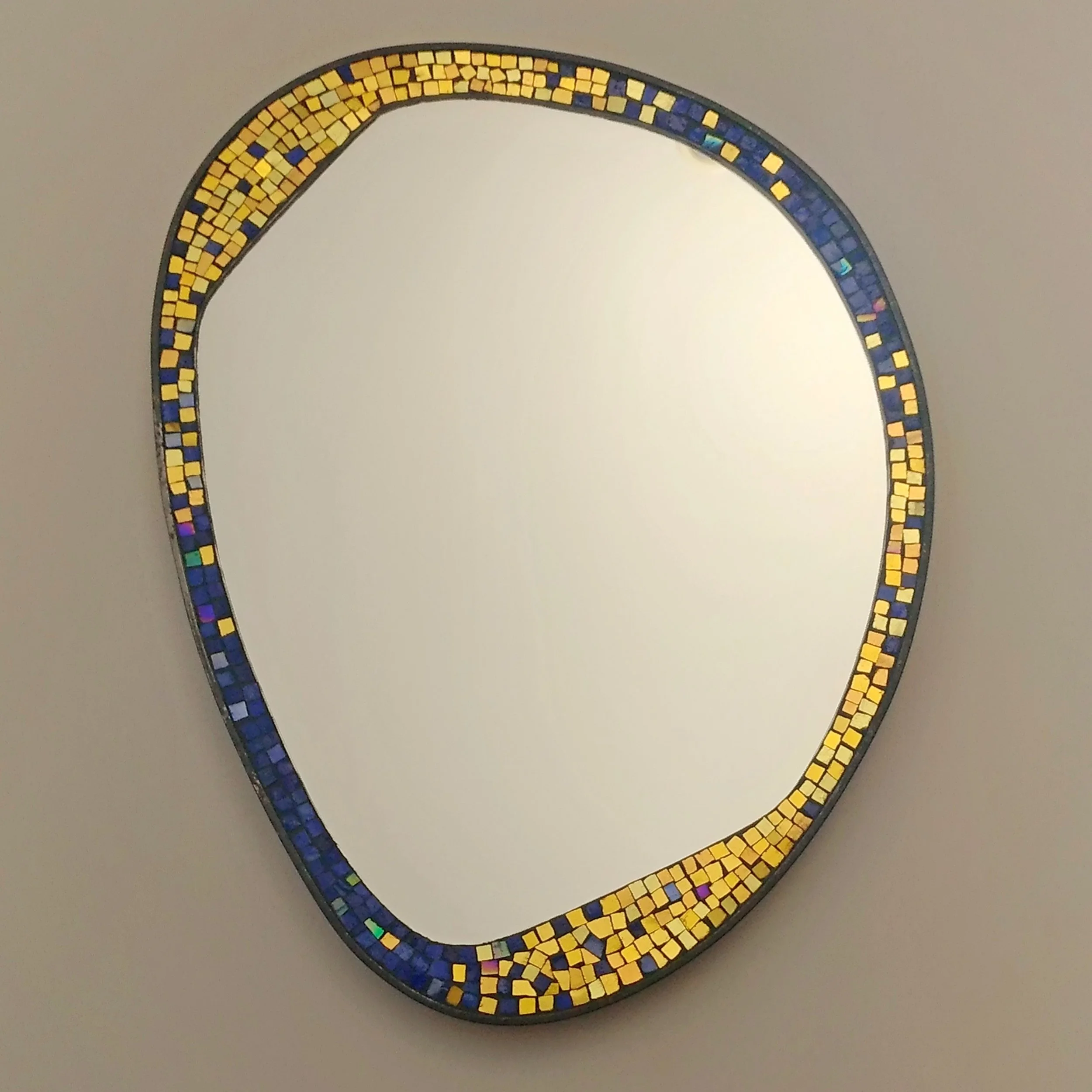 Miroir rond avec un cadre en mosaïque de petites tuiles colorées en jaune, bleu et violet, posé contre un mur clair.