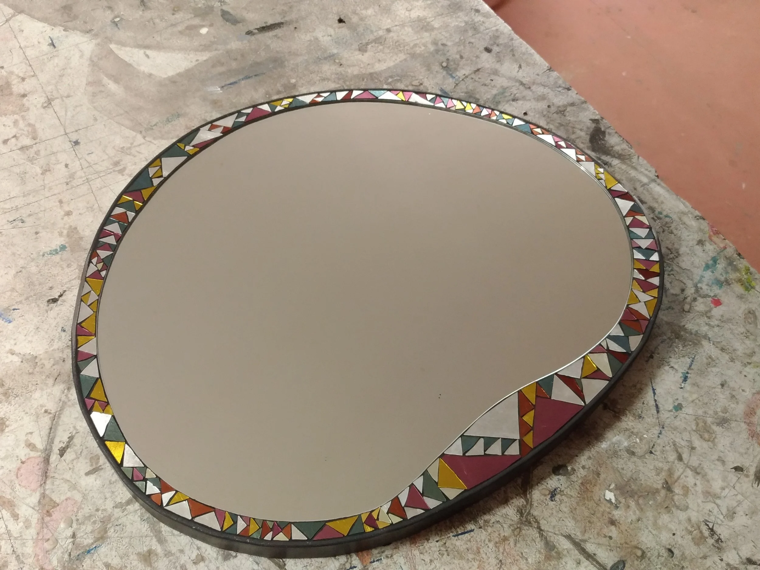 Un miroir en mosaïque rond avec un cadre décoratif en mosaïque de triangles colorés, posé sur un sol en béton avec des marques de peinture