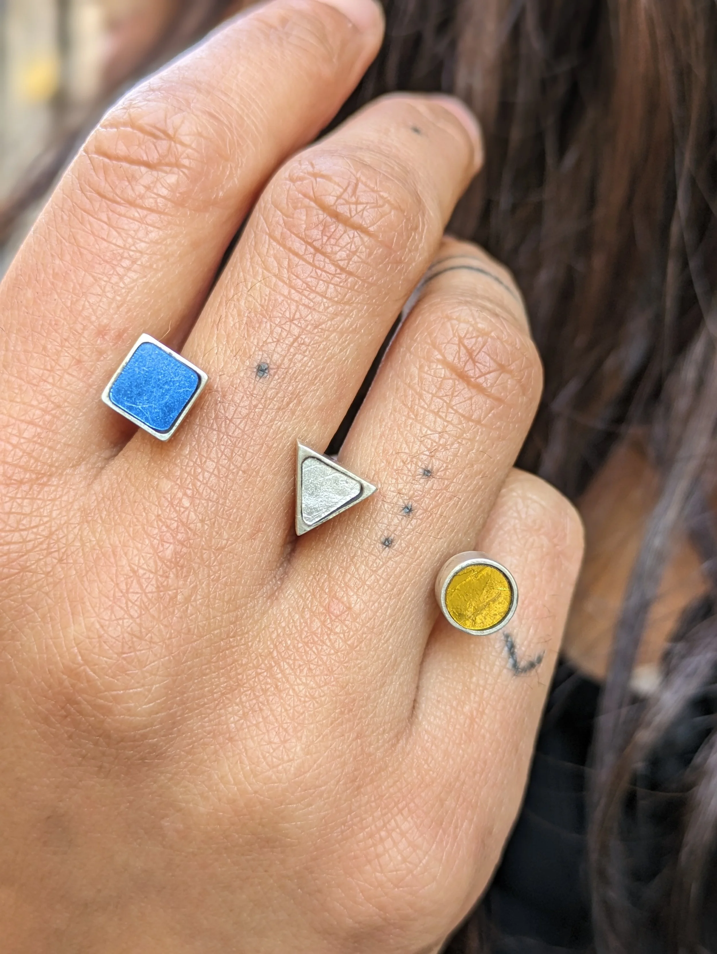 Une main nue portant une bague double en mosaïque de différents styles, dont une bague bleu clair, une bague argentée en forme de triangle, un anneau doré avec un disque jaune, sur le doigt avec la peau clair.