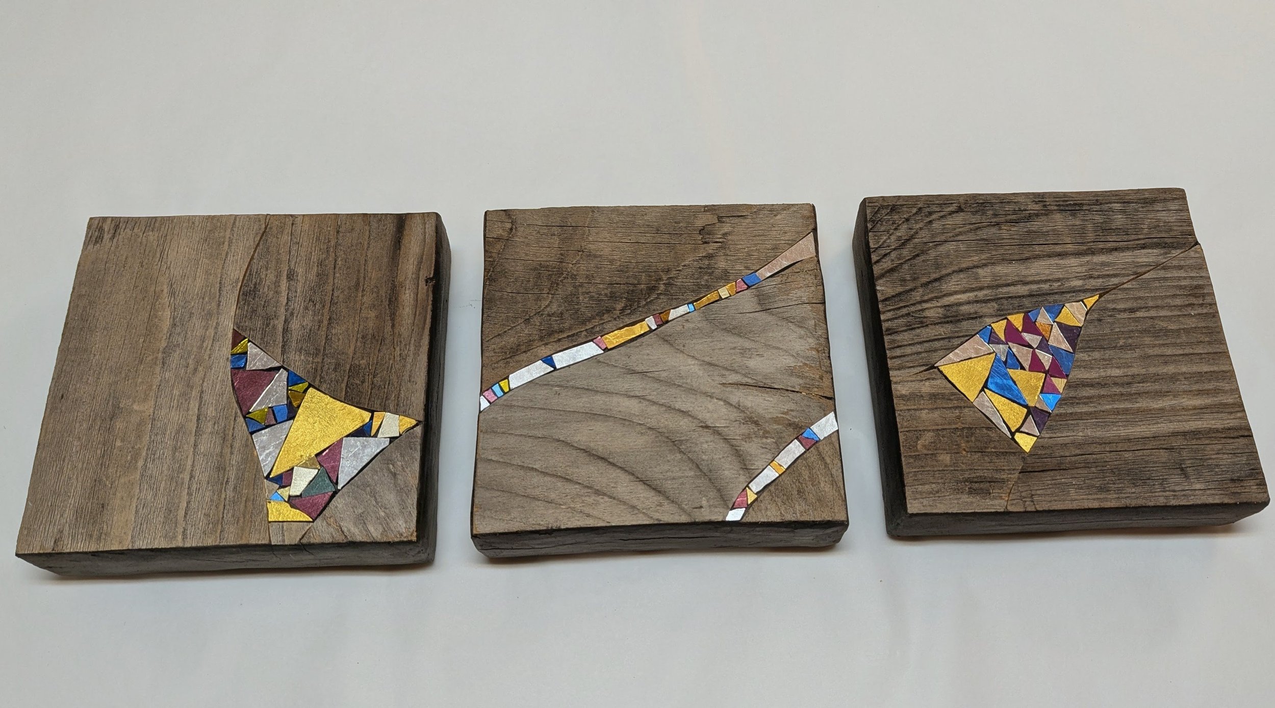 Trois morceaux de bois avec des mosaïques colorées en forme de triangles, formant un motif de flèche, sur un fond blanc.