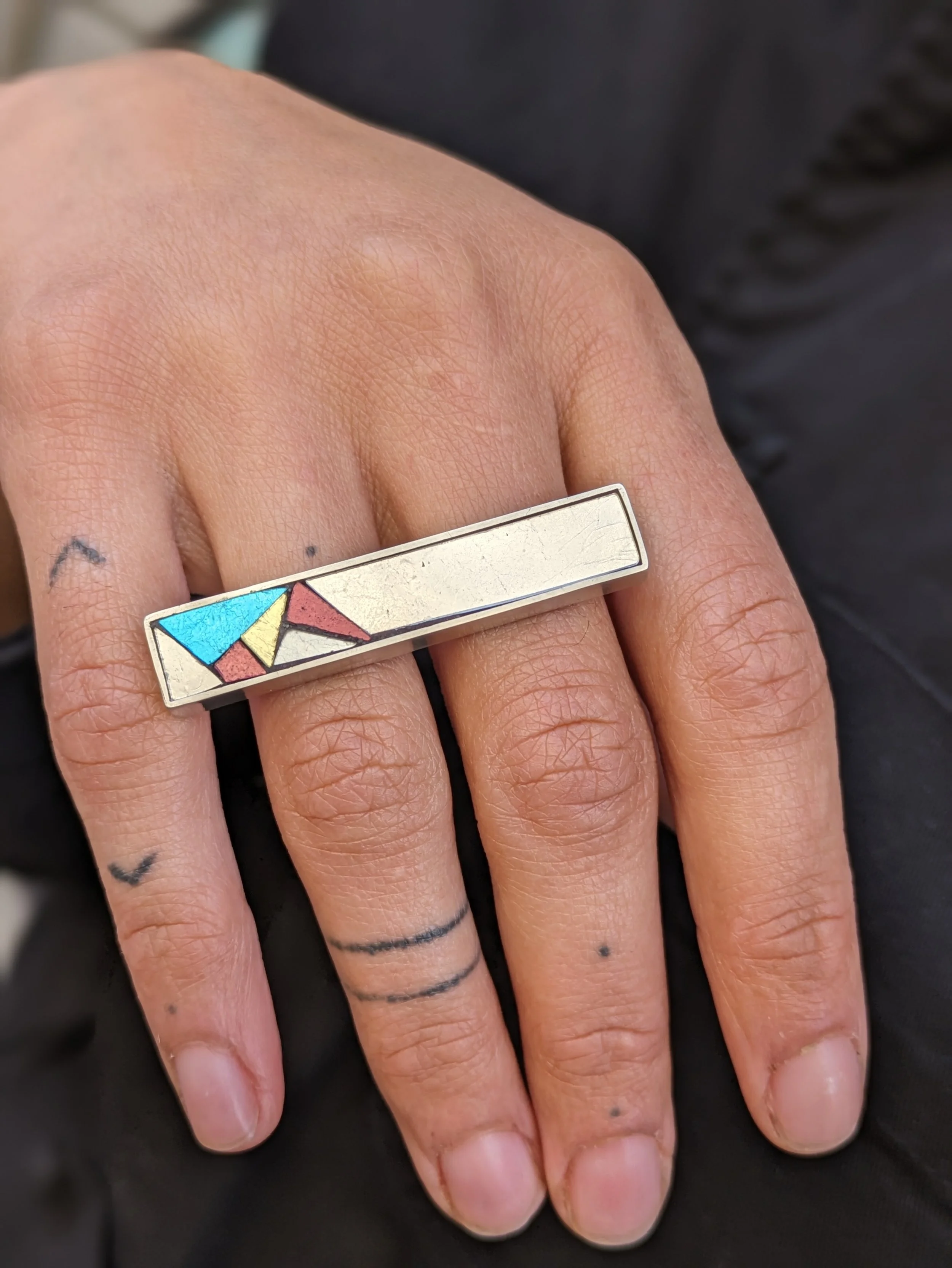 Main portant une bague rectangulaire en mosaïque avec un motif coloré abstrait.