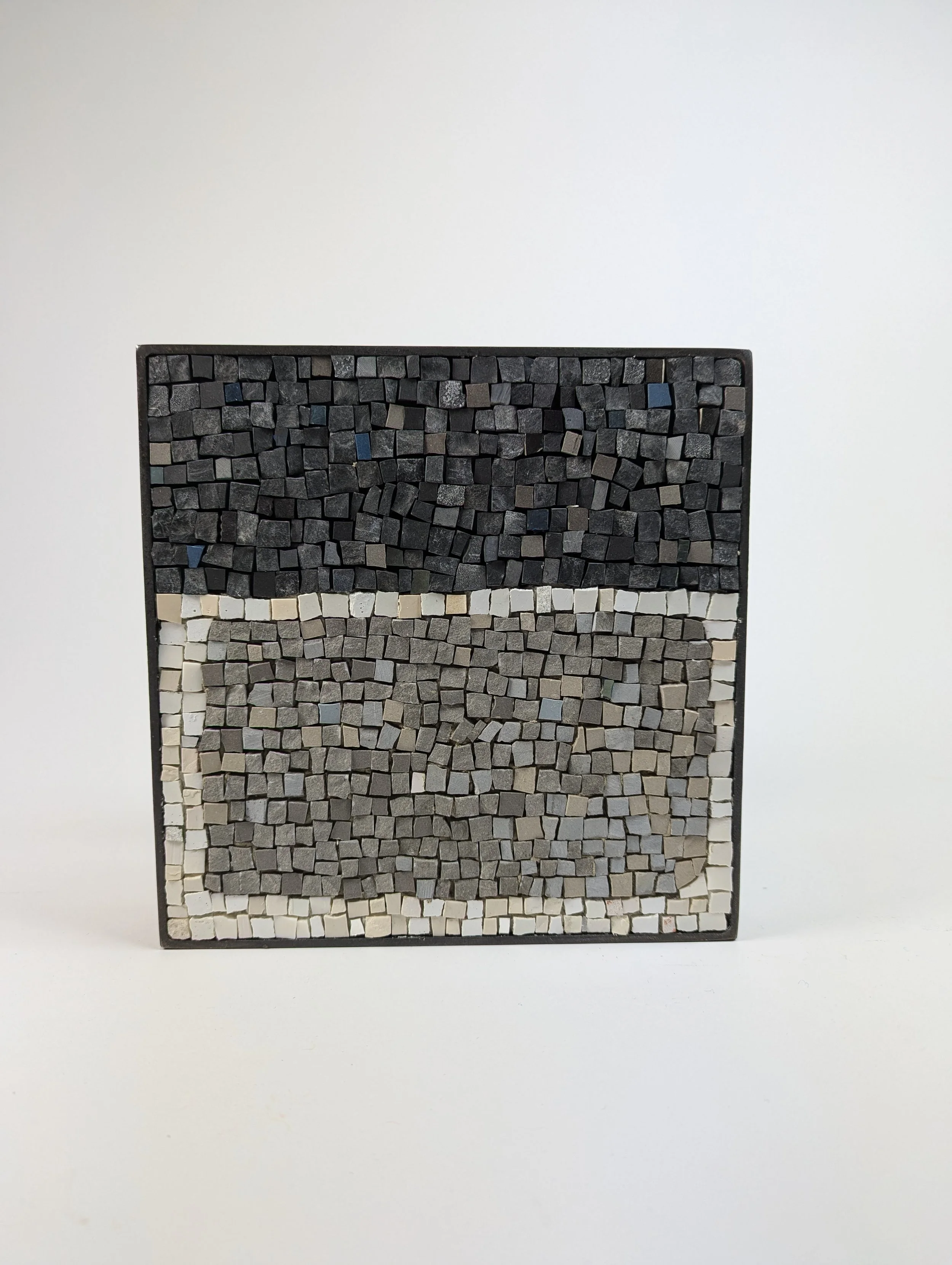Tableau en mosaïque de petits carrés en différentes teintes de gris, noir et blanc, dans un cadre noir, posé sur un fond blanc.