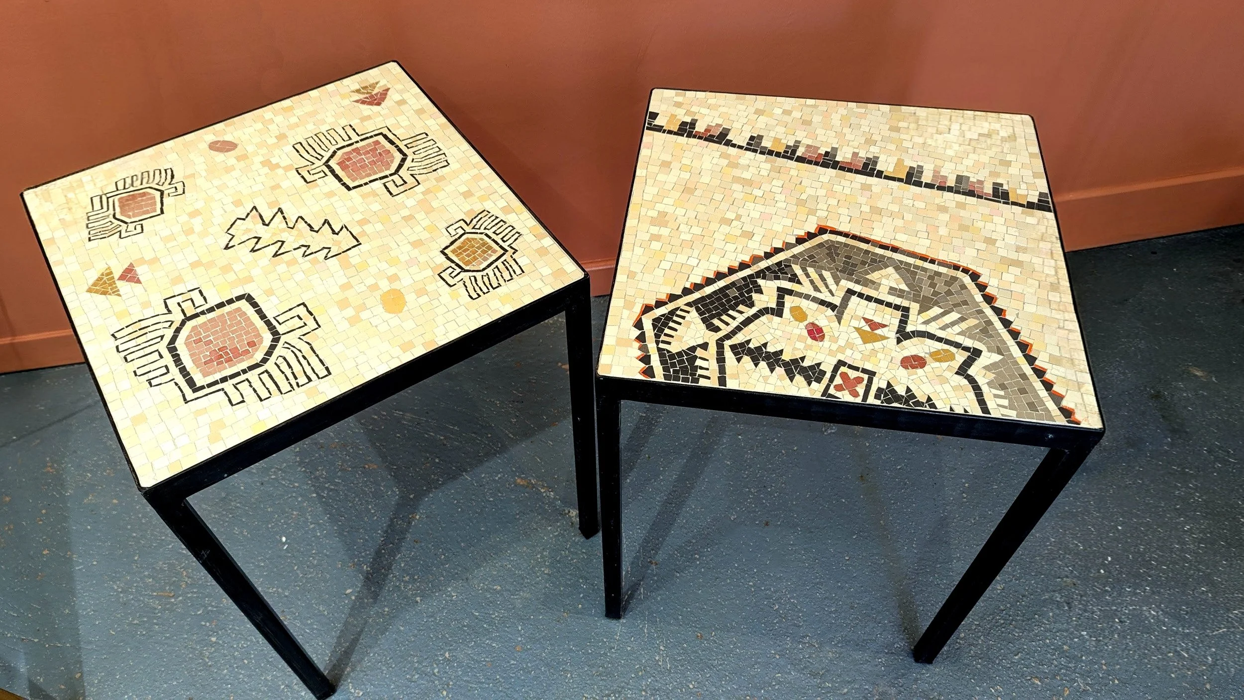 Deux tables en mosaïque avec des motifs géométriques et colorés, placées contre un mur de couleur beige ou rose clair.