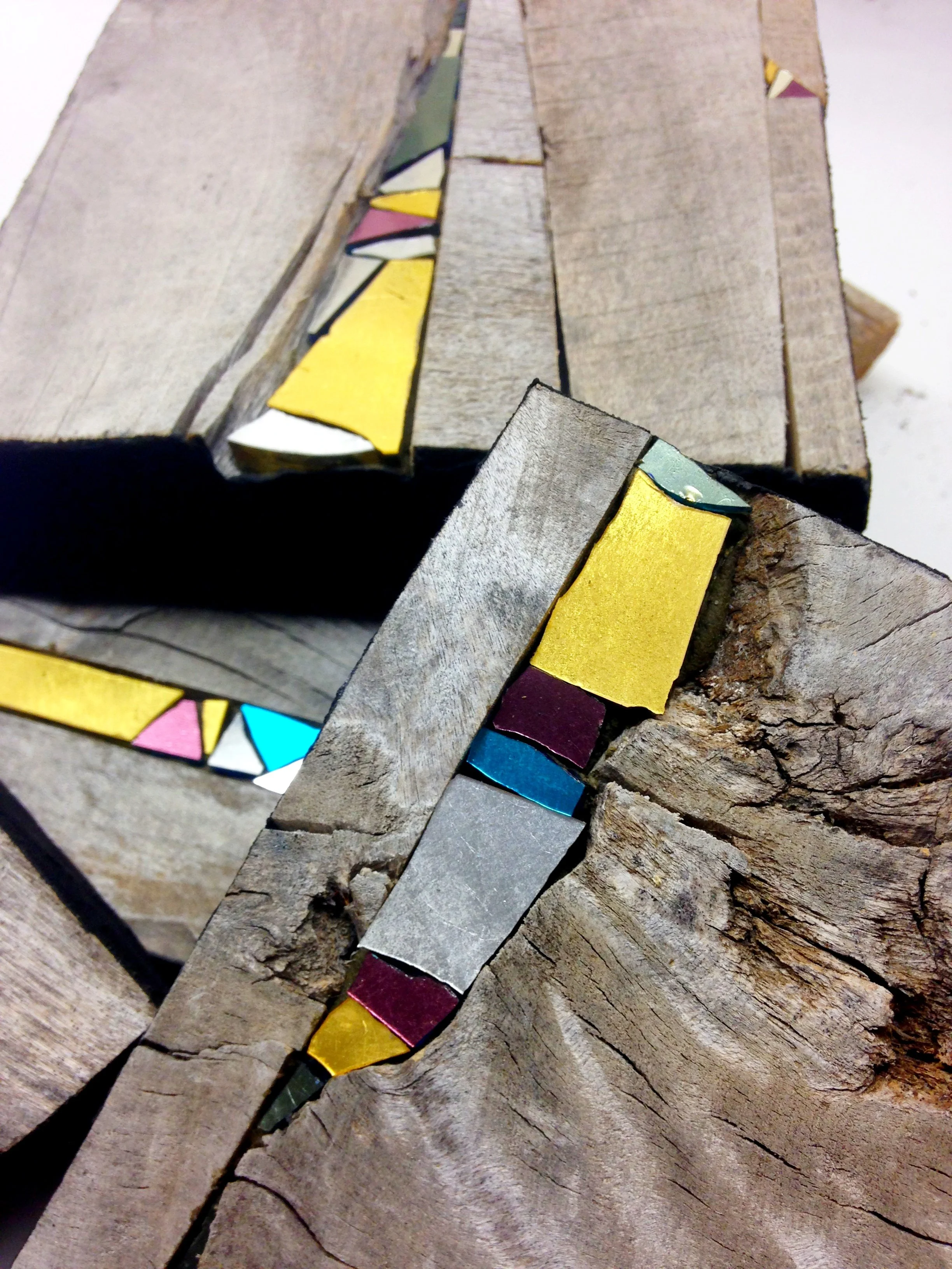 Bijou mural en mosaïque, un assemblage de morceaux de bois et de matériaux colorés, notamment jaune, vert, blanc, violet, bleu, et rouge, avec une texture de bois et des surfaces irrégulières.