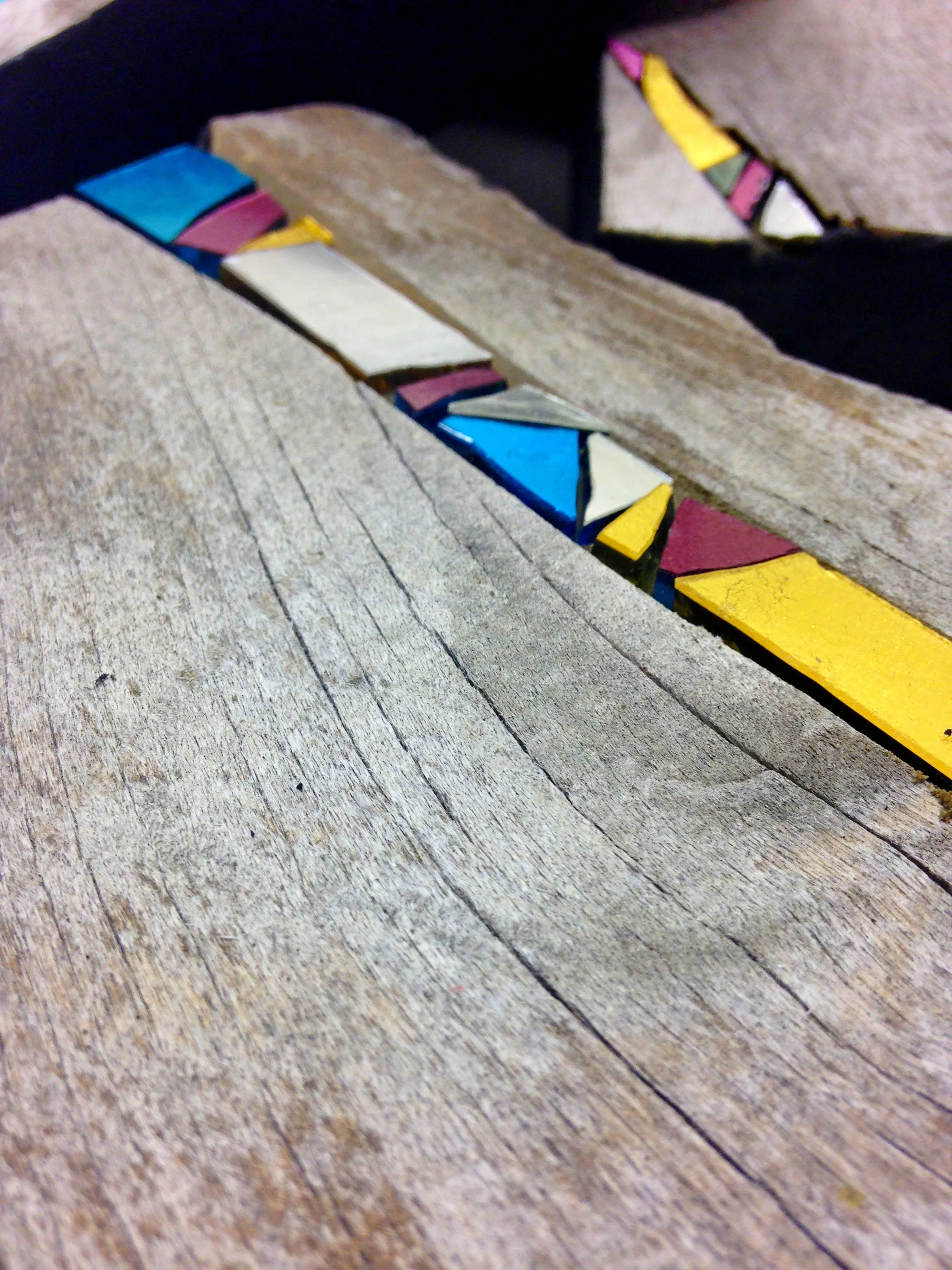 Bijou mural en mosaïque, surface en bois avec une rangée de carreaux colorés, principalement en bleu, jaune, rose, blanc, et noir, disposés le long d'une ligne.