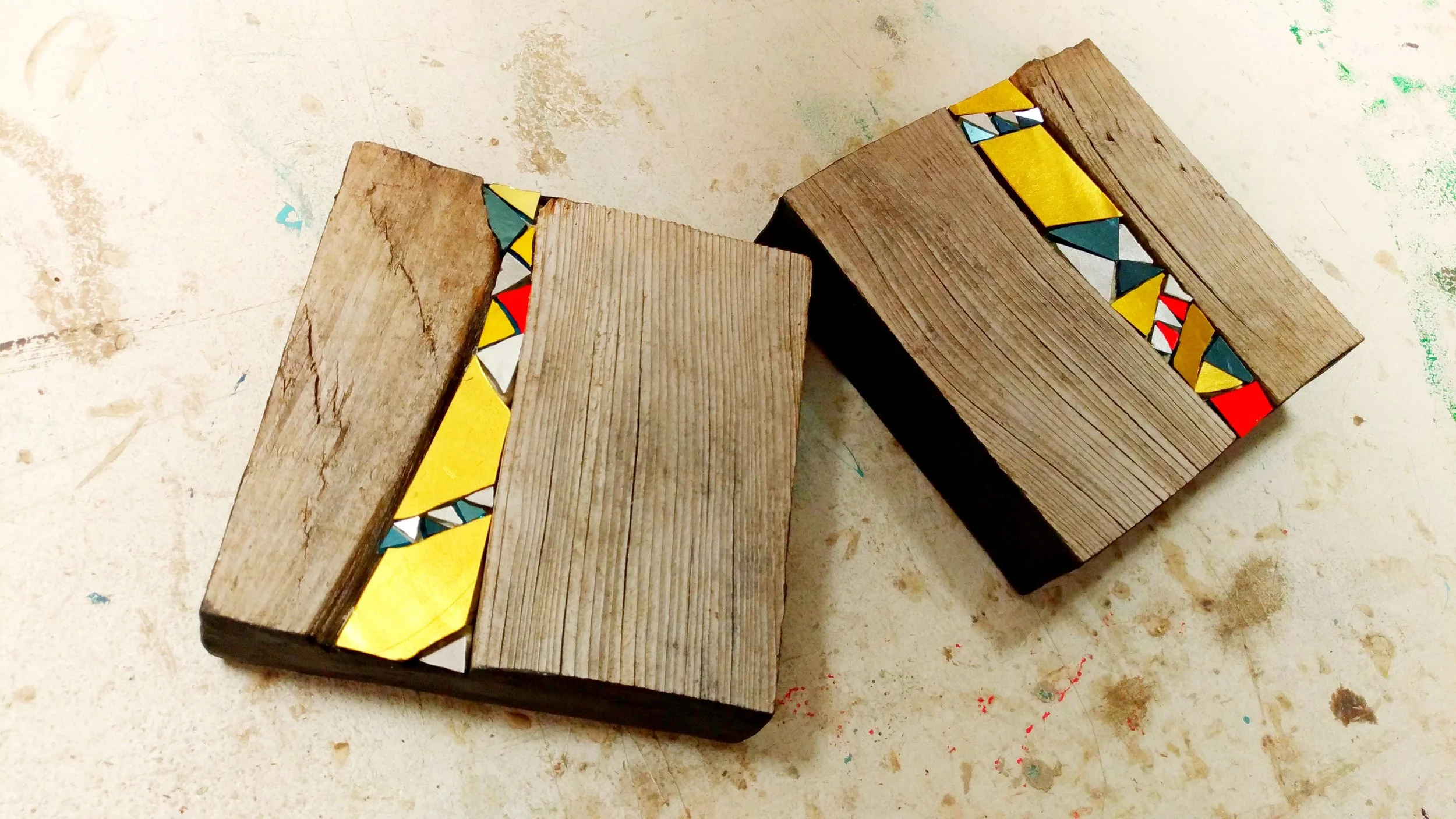 Trois blocs de bois décorés avec des morceaux de mosaïque colorée jaune, bleu, blanc et rouge posés sur une surface poussiéreuse ou tachée.