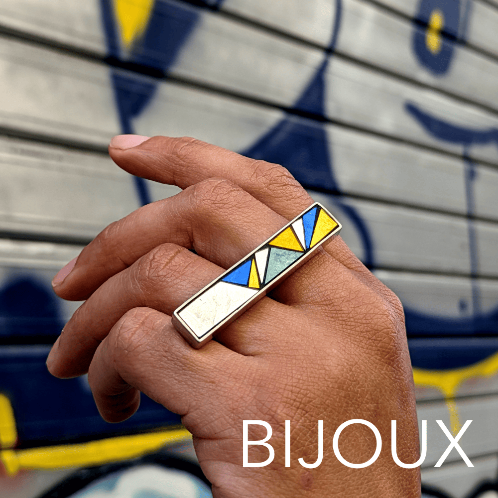 Une main porte une bague en mosaïque avec un design géométrique coloré, en arrière-plan un mur en bois peint avec du grafiti.