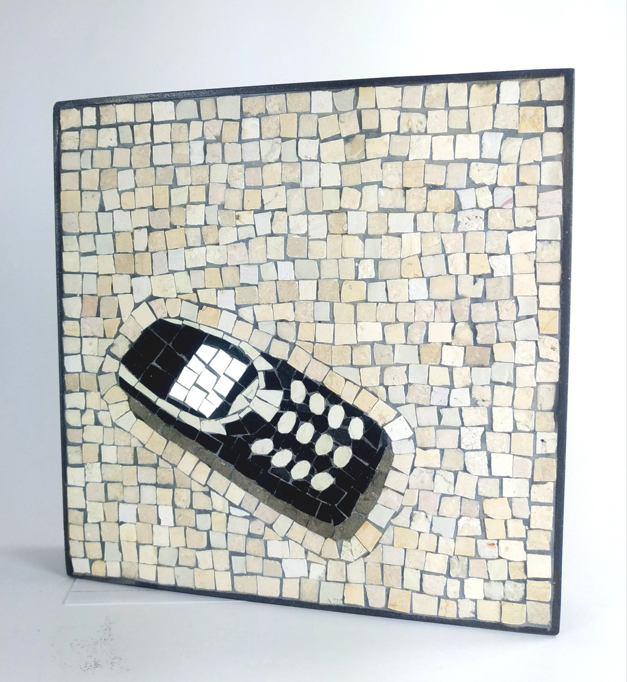 Tableau en mosaïque décorative représentant un téléphone portable, composée de petites pierres noires et blanches, encadrée par une bordure noire, installée sur un mur en pierre beige.