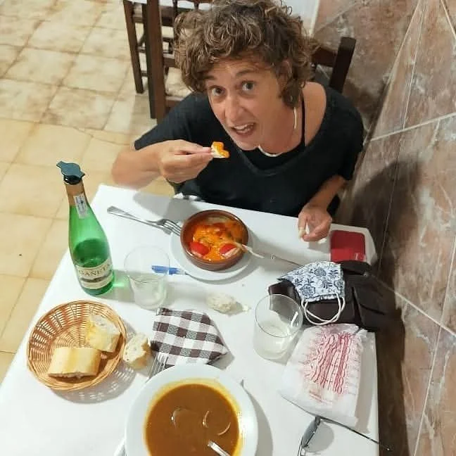 Une femme souriante mange une cuillère de nourriture dans un restaurant. La table est dressée avec une bouteille de boisson gazeuse, un bol de soupe, un panier de pain, un verre d'eau, une nappe en tissu, et d'autres objets.