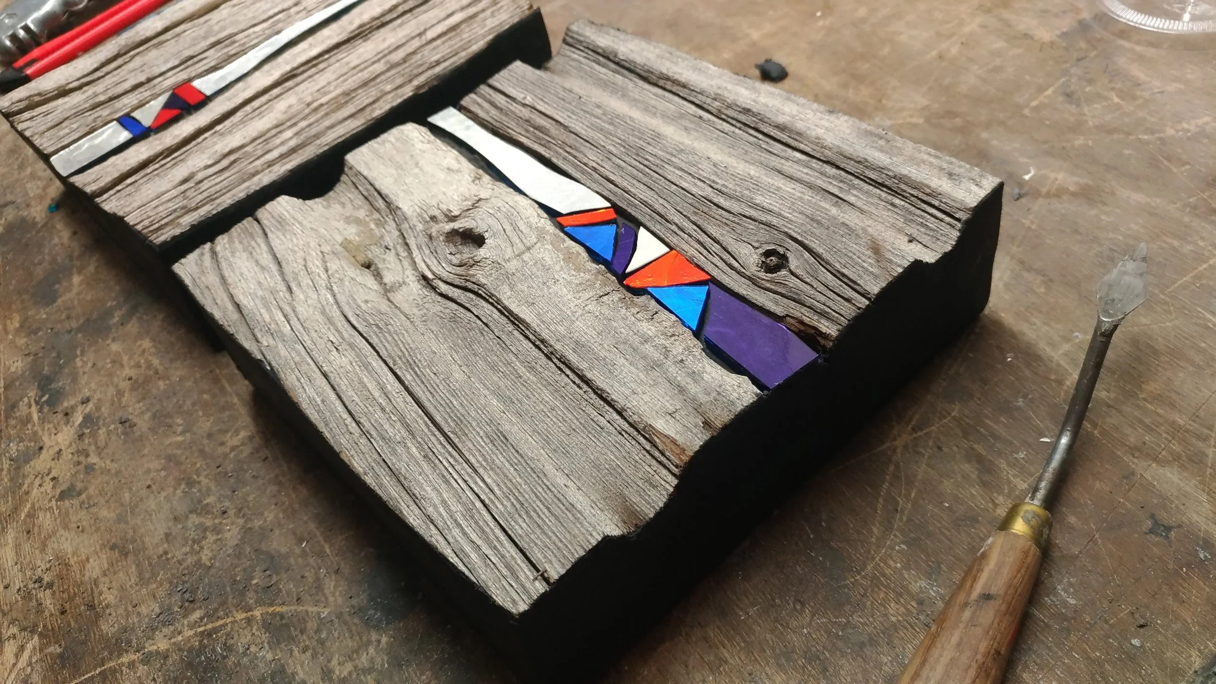Un morceau de bois ancien avec des morceaux de verre colorés en mosaïque insérés, associé à un outil en métal à la poignée en bois sur une surface de travail en bois.