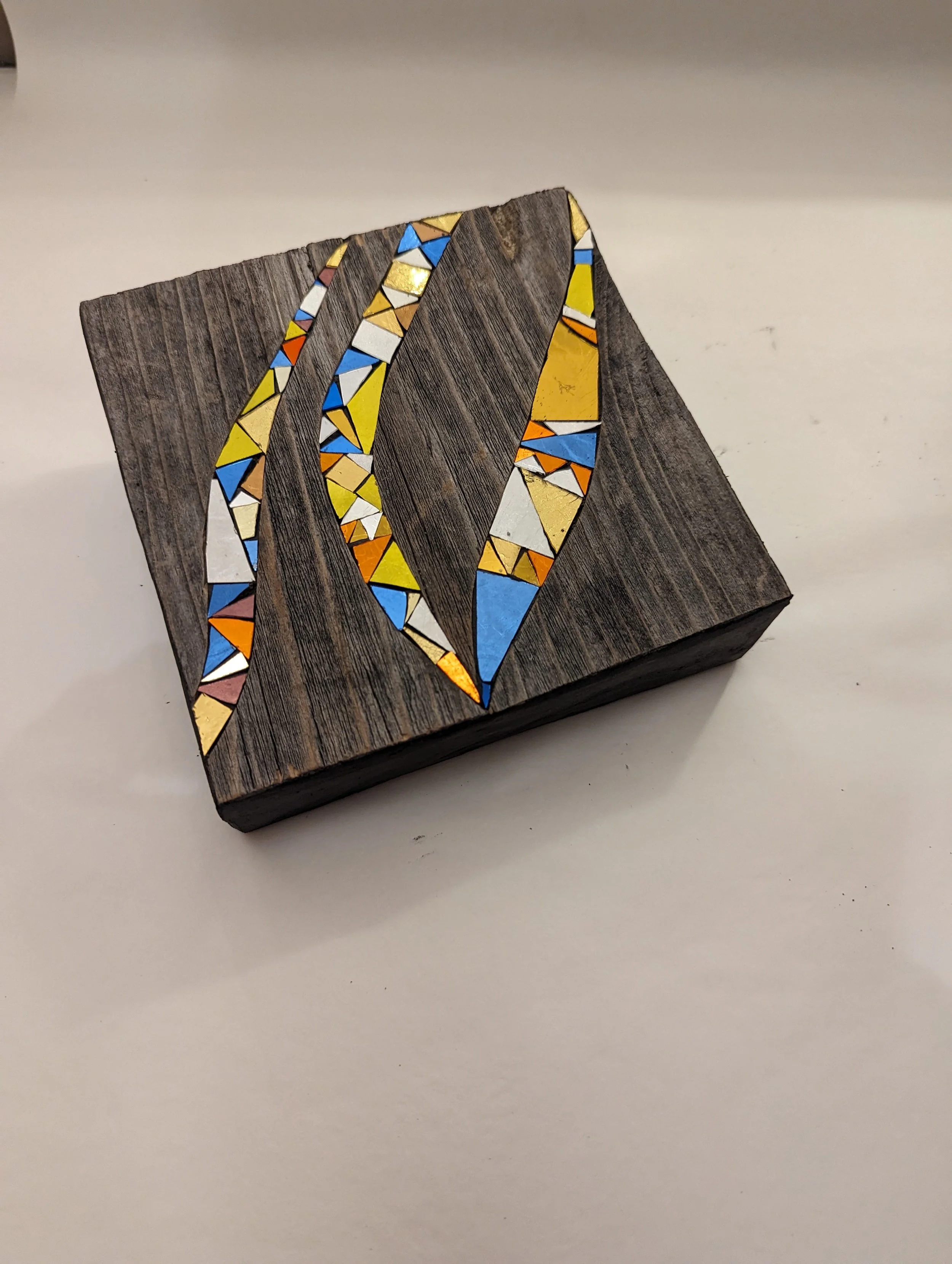 Une pièce en bois avec un motif de mosaïque colorée, comprenant des morceaux de verre ou de céramique jaune, blanc, bleu, orange et rouge, formant des lignes ondulantes.