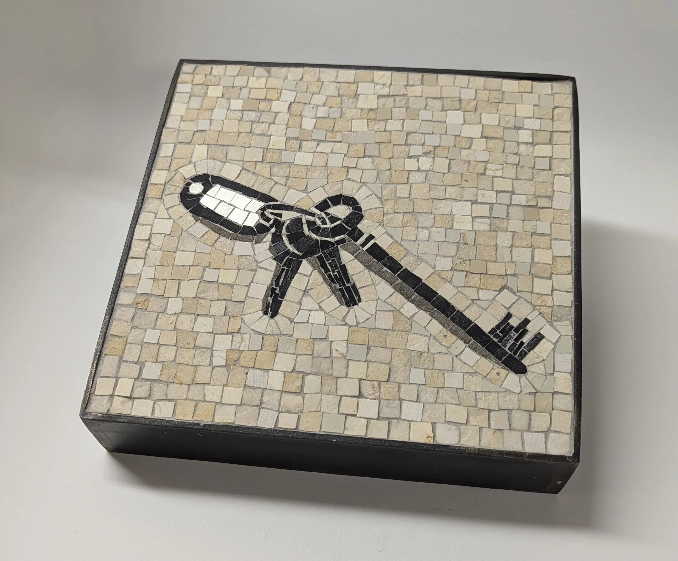 Une mosaïque représentant une clé en noir et blanc, entourée de tesselles de pierre beige, encadrée par une bordure noire.
