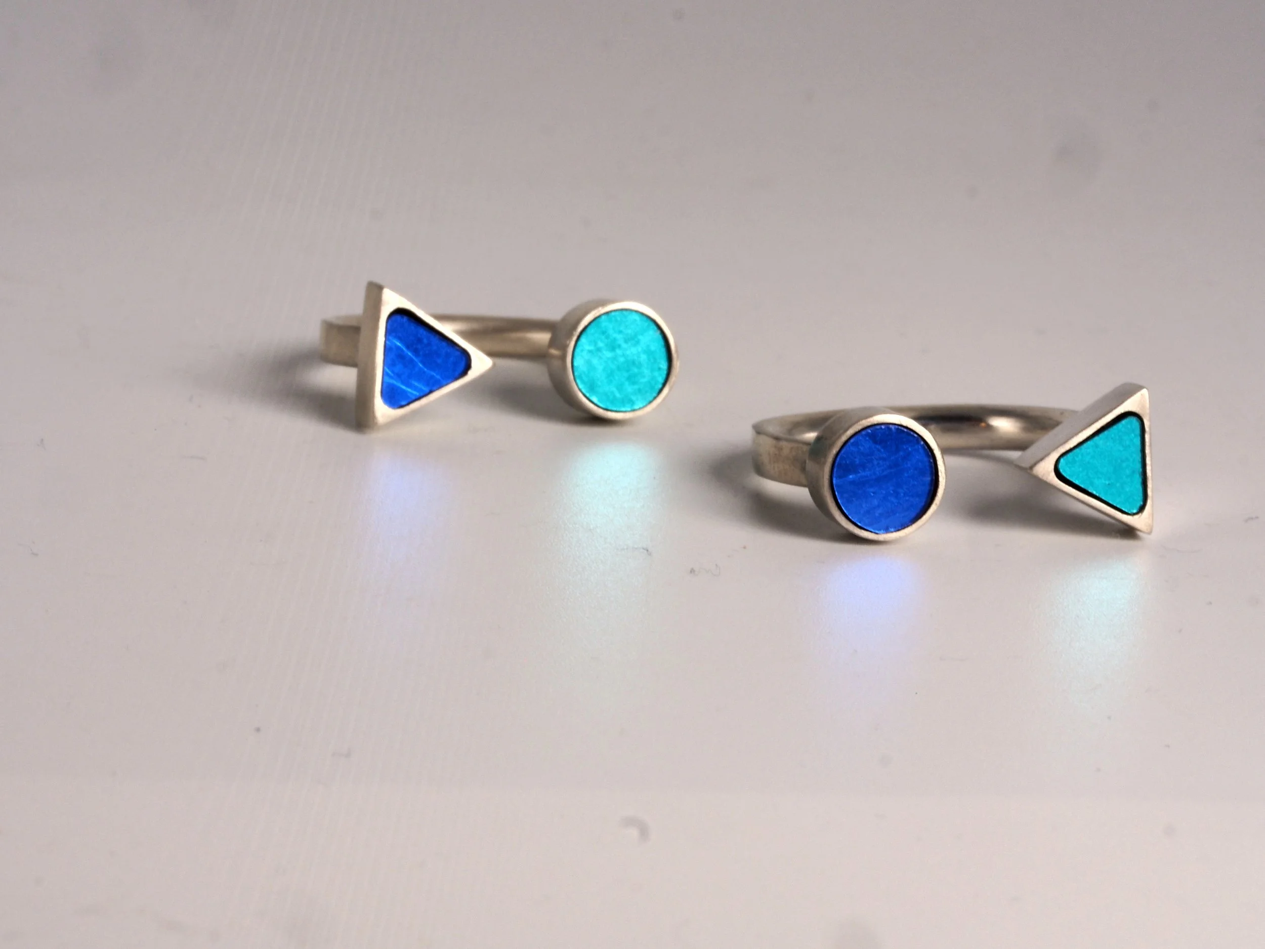 Bague en mosaïque en forme de triangles et cercles colorés en bleu et turquoise posés sur une surface blanche.