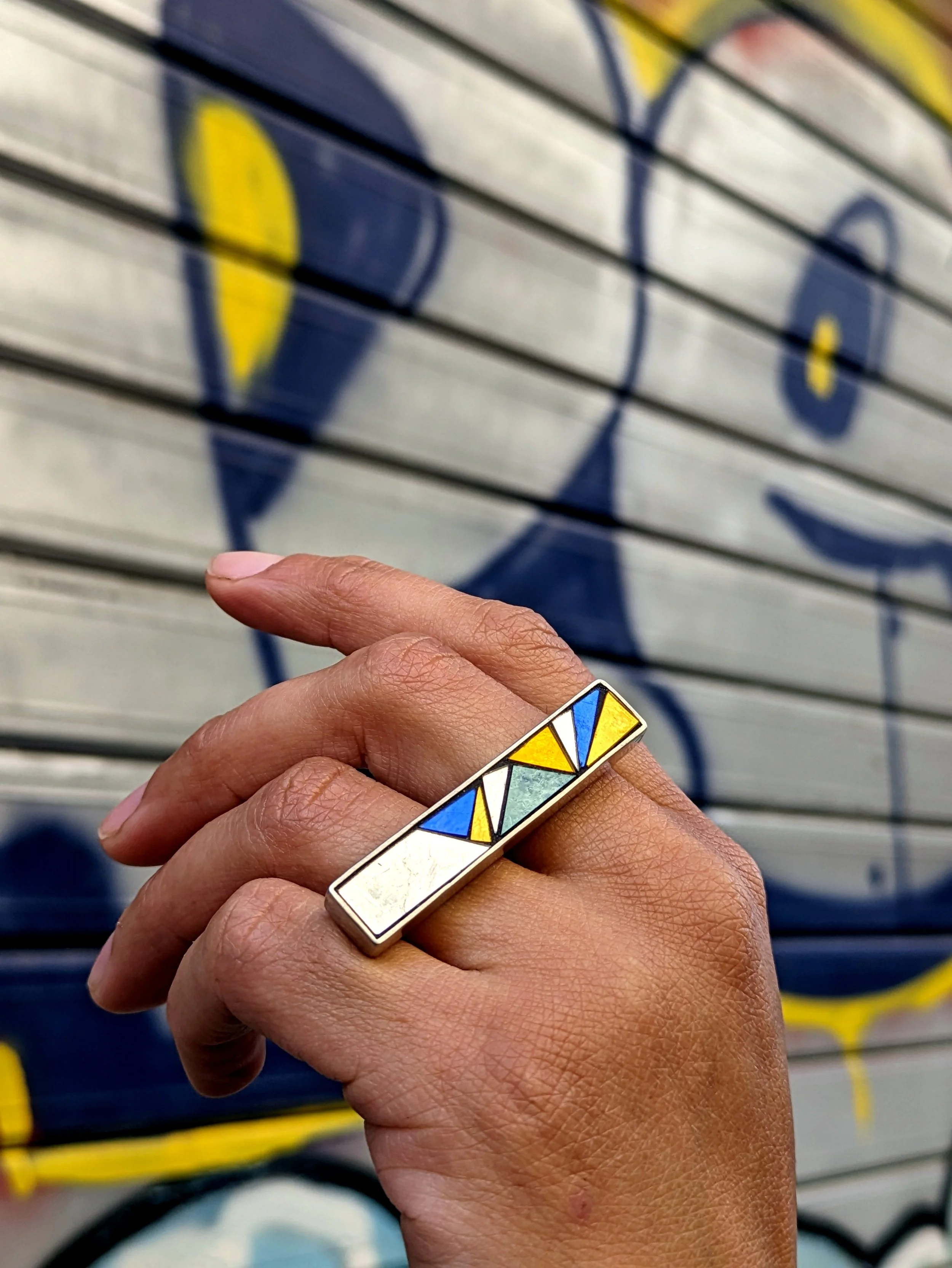 Une main tient une bague en mosaïque avec un design multicolore. En arrière-plan, un mur avec des graffitis bleus et jaunes.
