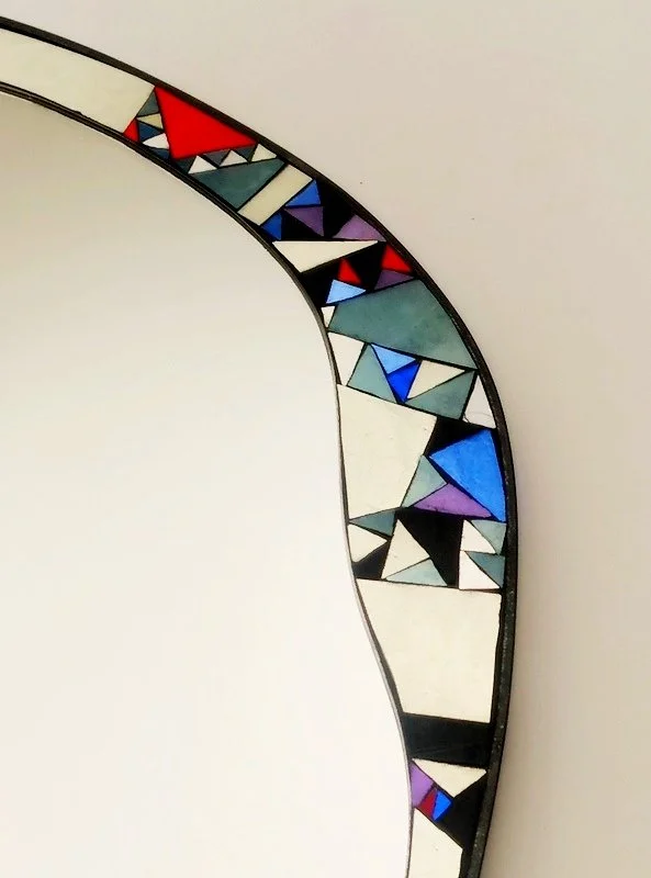 Un miroir en mosaïque coloré avec des motifs géométriques dans un cadre en métal, formant une courbe partielle sur un mur blanc.
