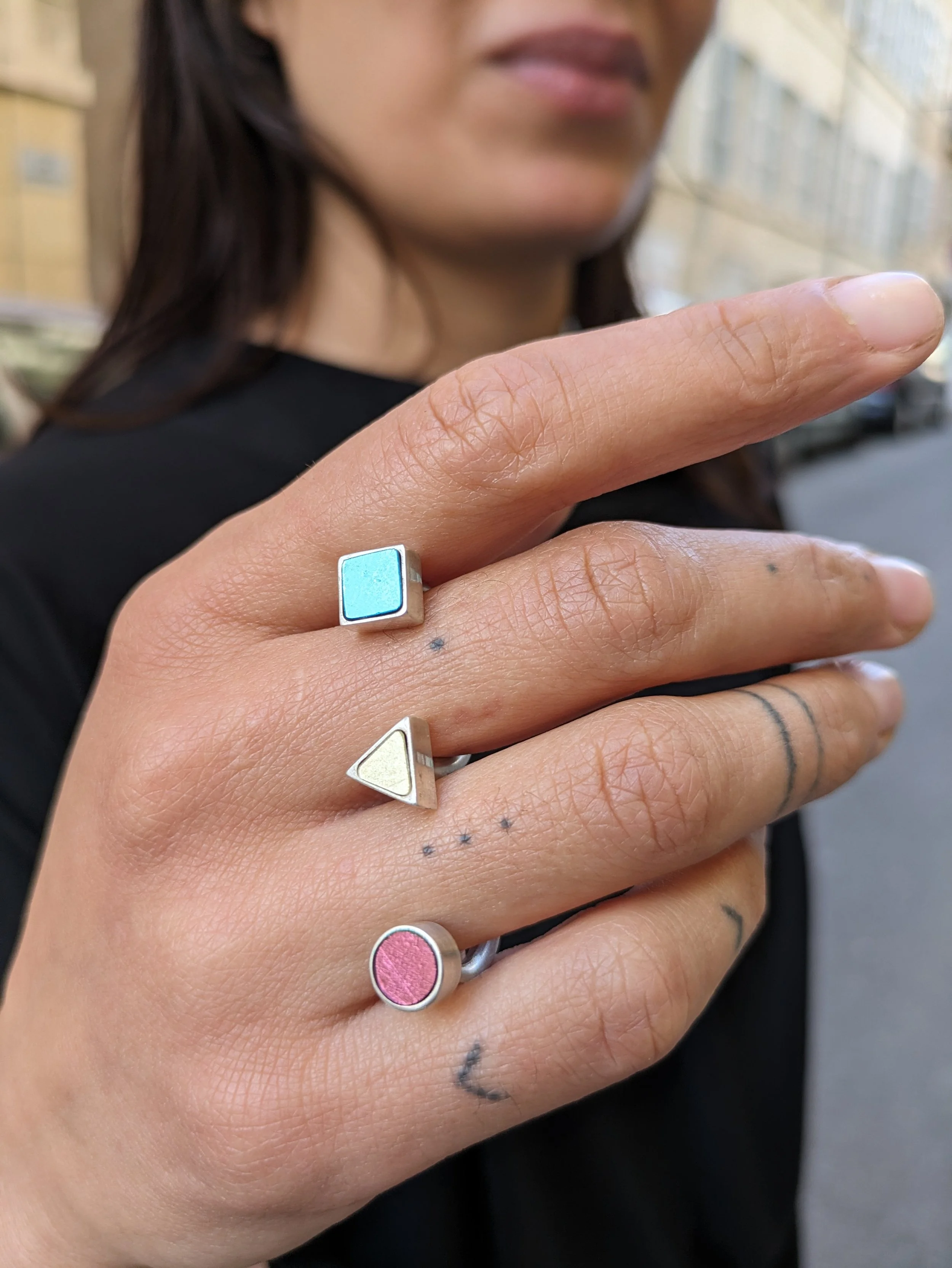 Une main féminine porte une bague double en mosaïque, avec un fond flou montrant une femme et un quartier urbain.