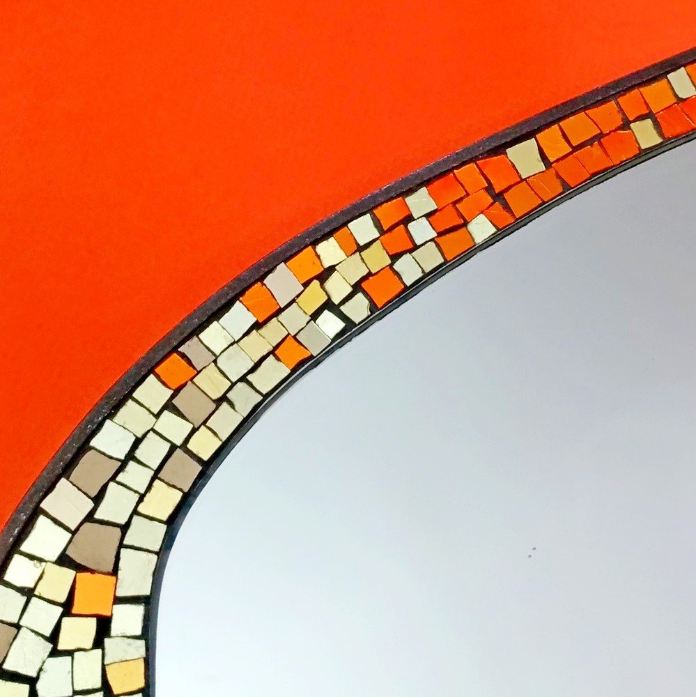Détail d'une surface en miroir avec un cadre orné de petits carreaux de mosaic aux couleurs ivoire, beige, orange et noir, contre un fond rouge.