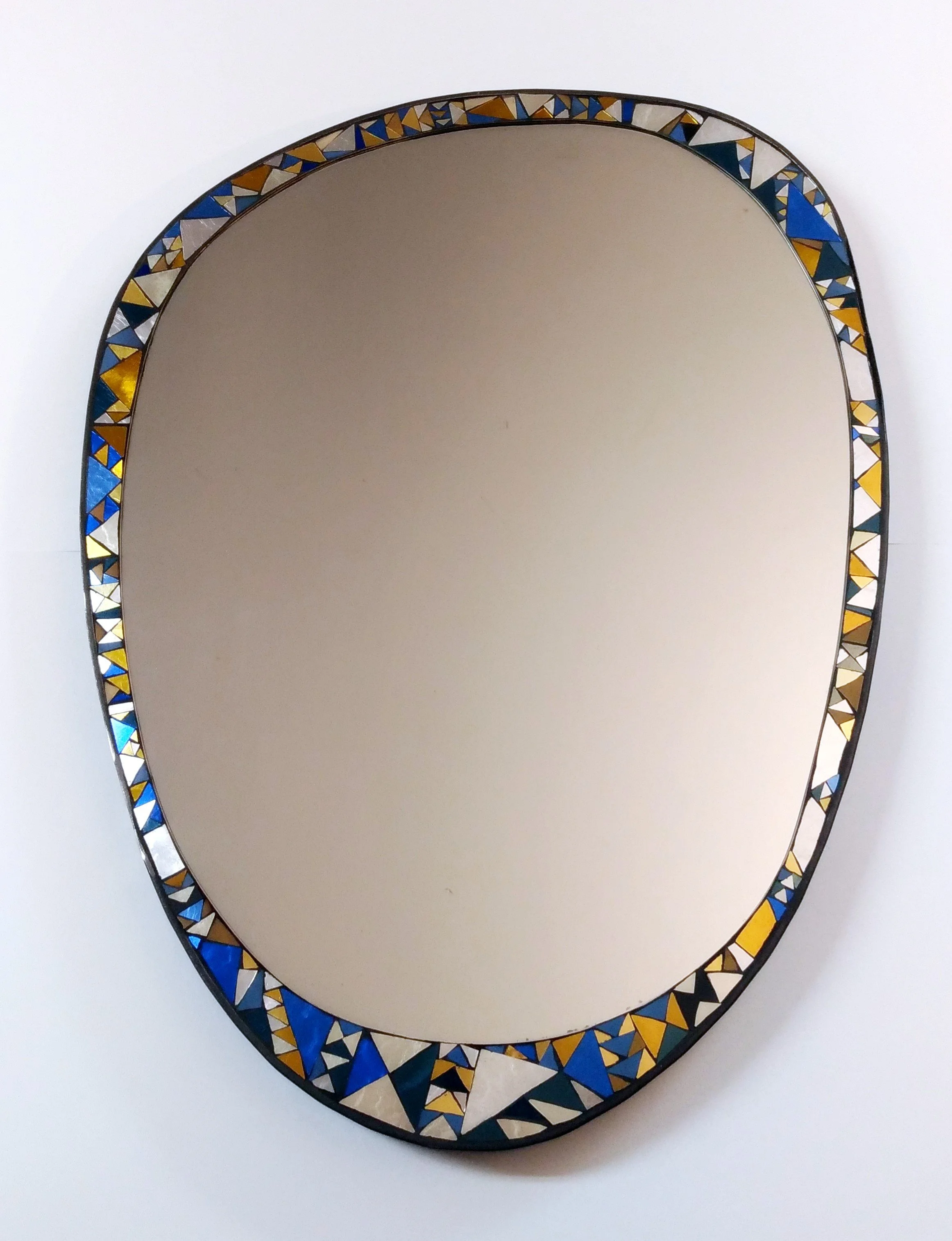 Miroir ovale avec cadre en mosaïque de triangles colorés en bleu, or, blanc, noir et beige, posé sur un fond blanc.