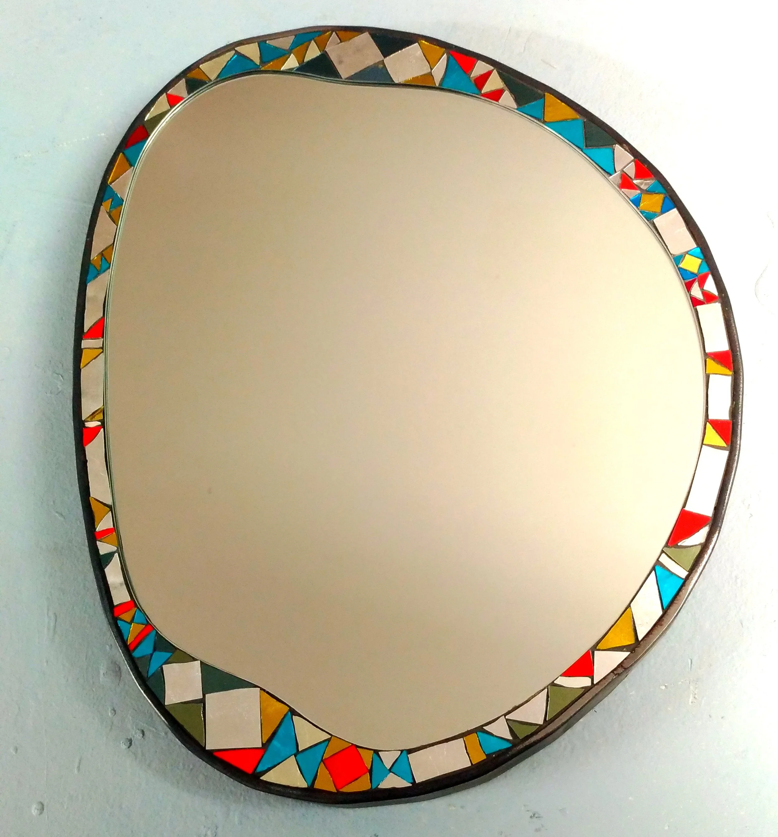 Miroir de forme ovale en mosaïque avec un cadre en mosaïque colorée, posé sur une surface gris clair.