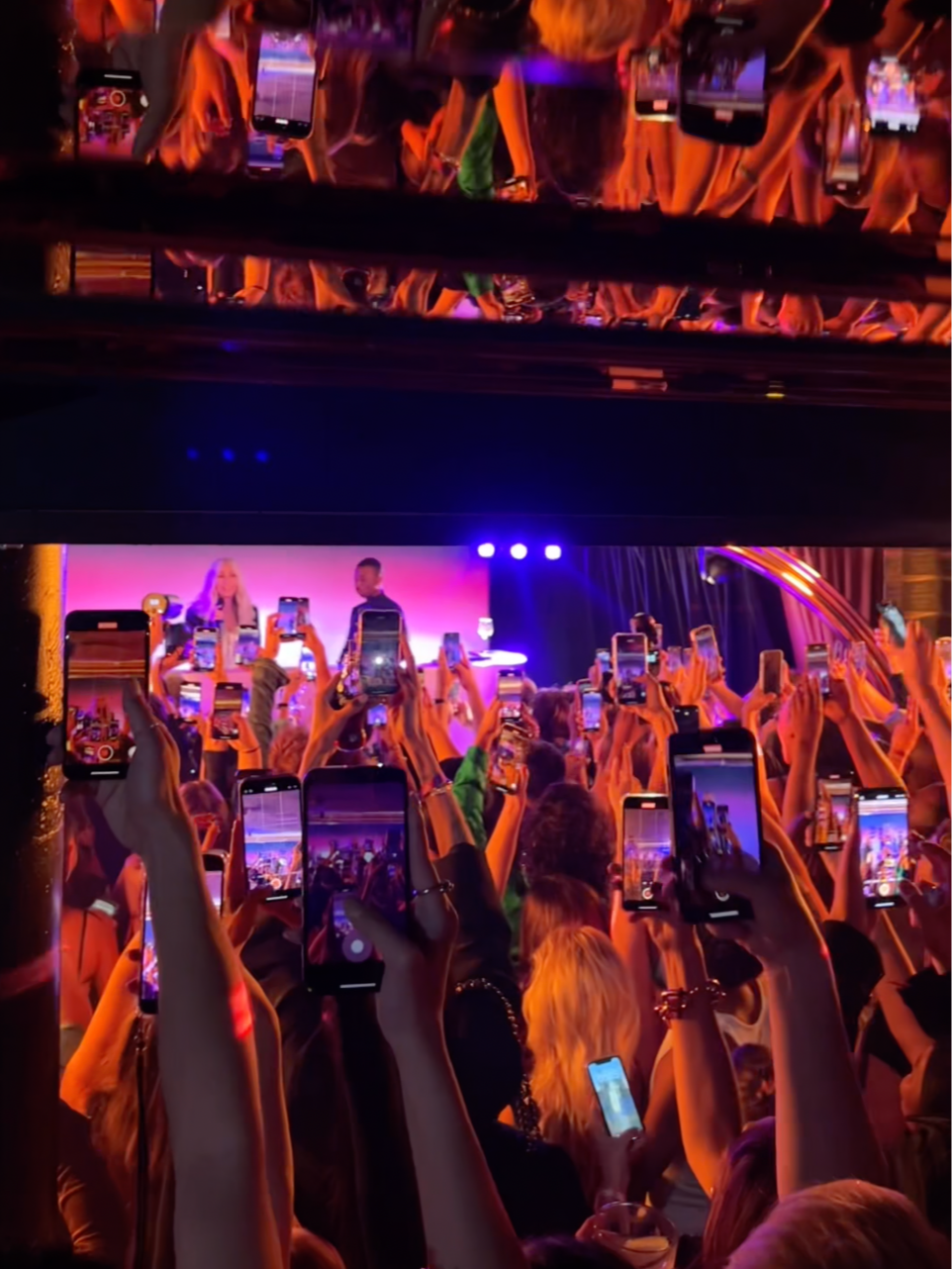Crowd at a concert or event, many people are holding up smartphones to take photos or videos of two performers on stage with pink and purple lighting.