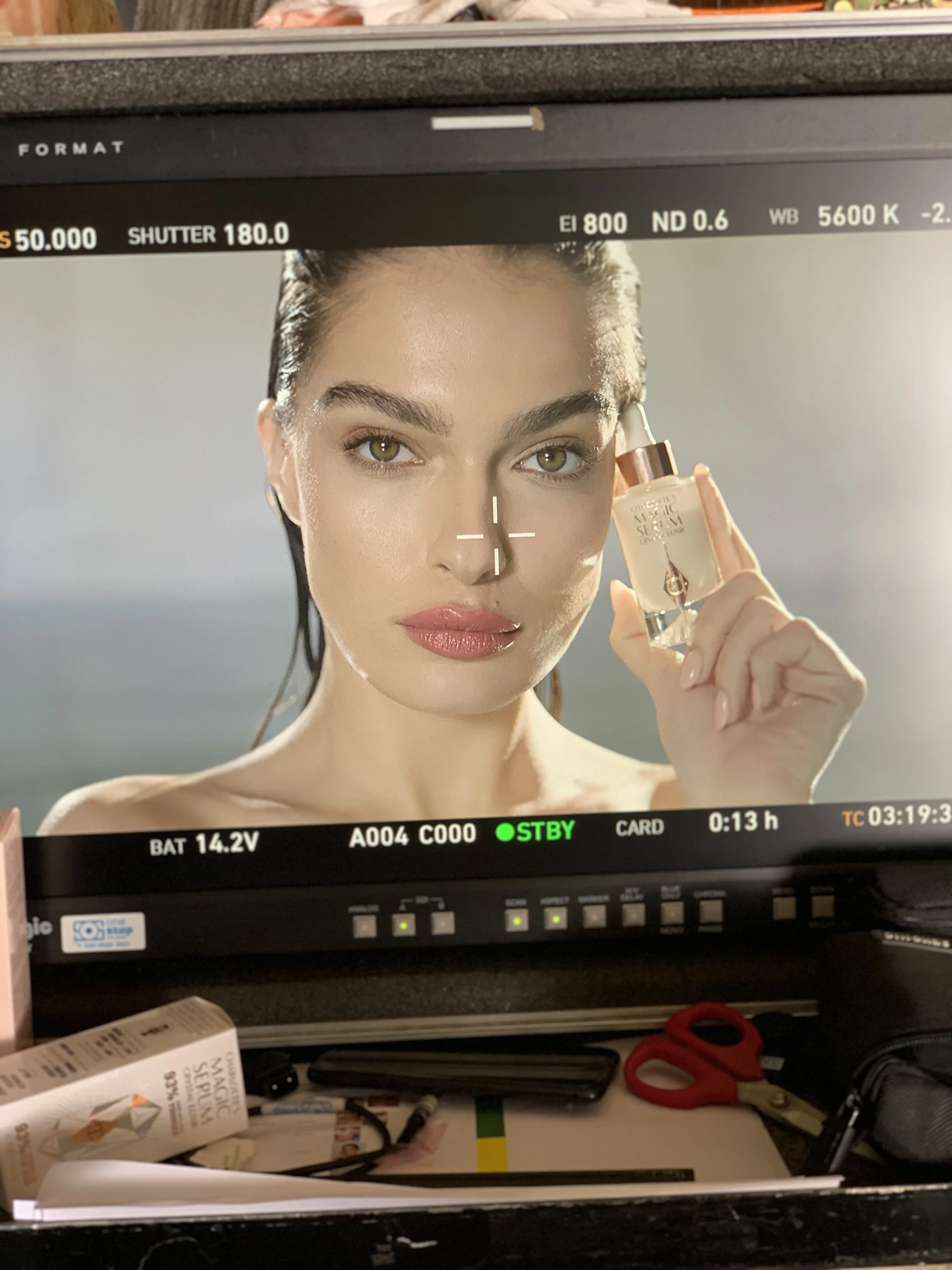 The image shows a monitor displaying a close-up of a woman holding a small bottle of product near her face, with makeup and a focused expression.