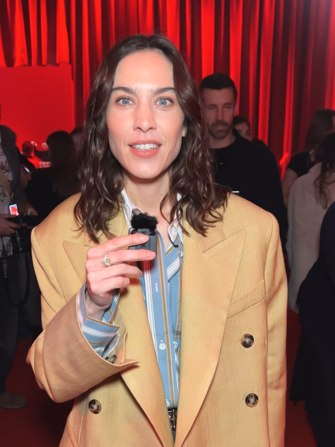 Alexa Chung wearing a beige blazer and a colorful blouse holds a microphone and smiles at the camera at an indoor event with red curtains and other people in the background.
