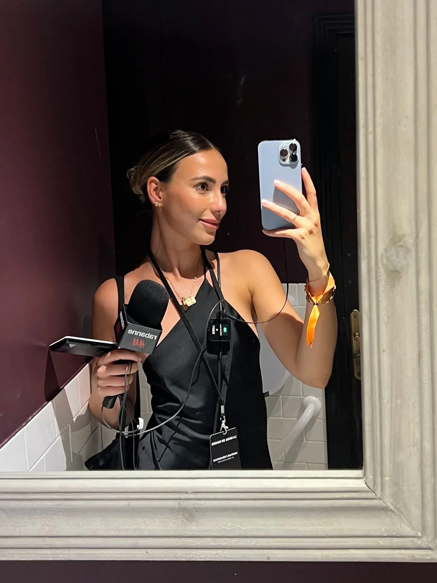 Marcia taking a selfie wearing a black sleeveless top and has a name tag. She is holding a microphone and a recording device. The setting appears to be a bathroom with tiled walls and a door handle visible.