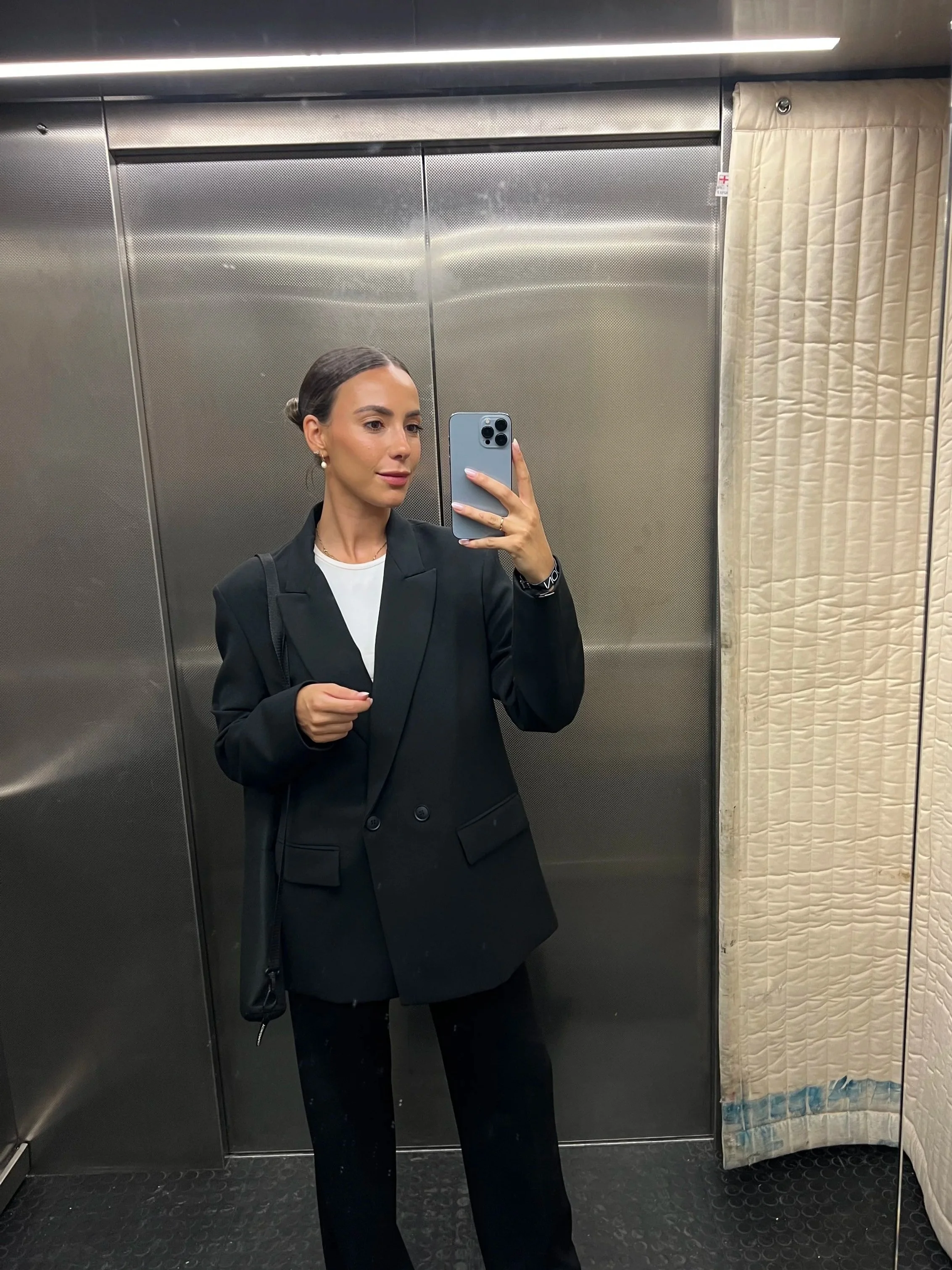 Marcia inside an elevator. Wearing a black blazer over a white top, and holding a phone in her right hand. There is a beige curtain on the right side of the elevator.