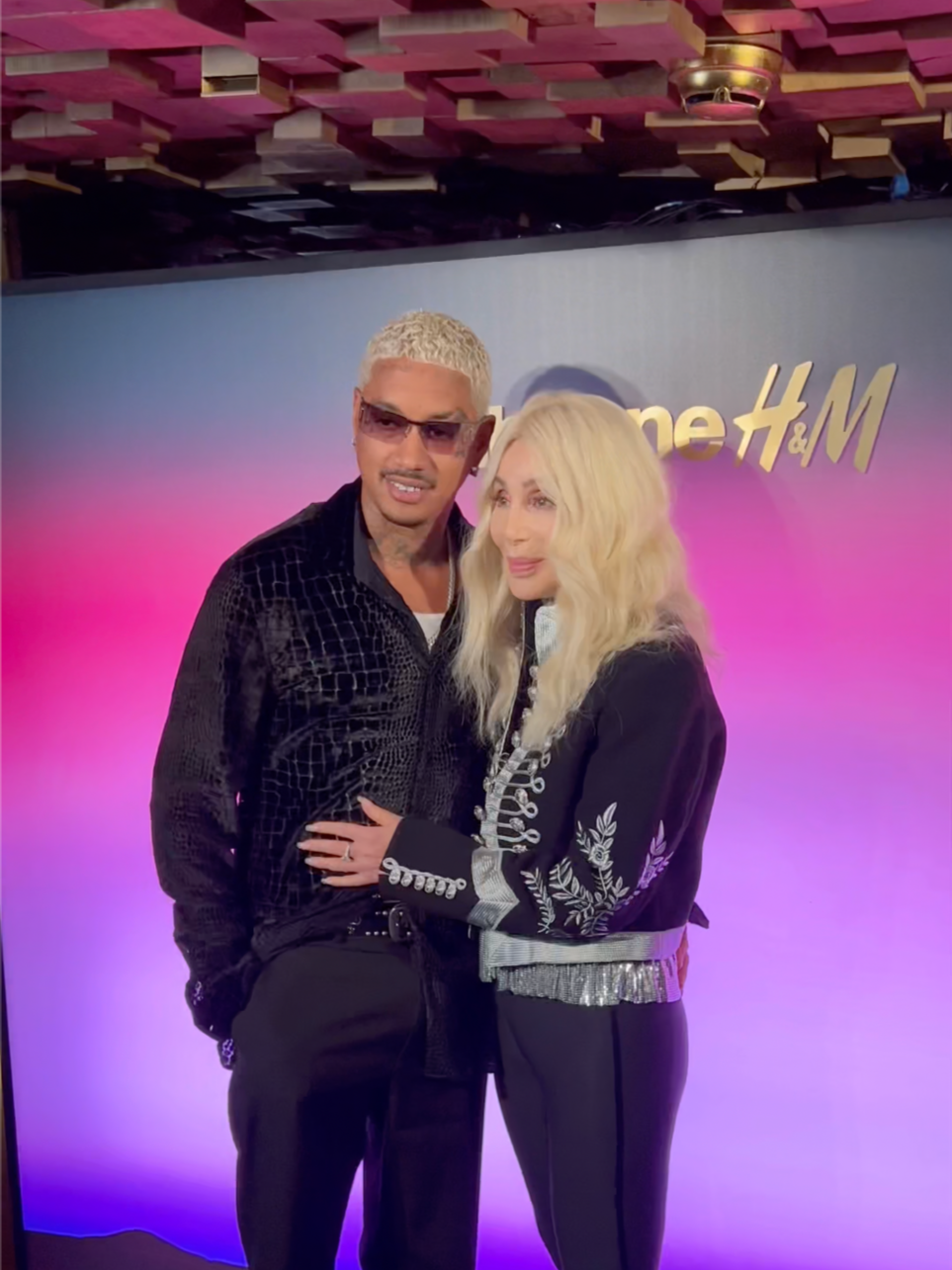 Cher and her boyfriend Alexander "A.E." Edwards standing together in front of an H&M backdrop, with pink and purple lighting in the background.