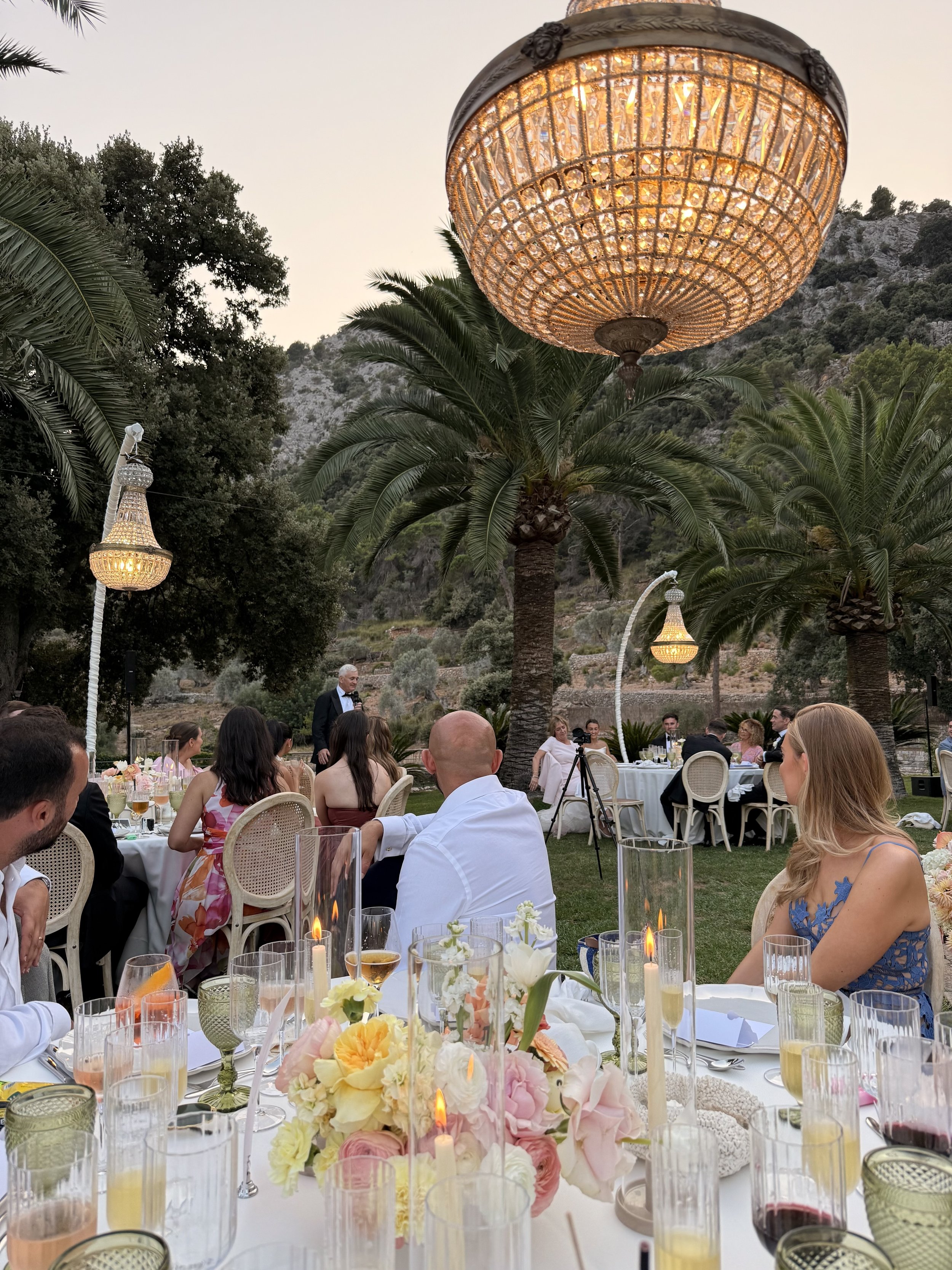 Outdoor wedding reception in Mallorca under chandeliers with palm trees and mountain views