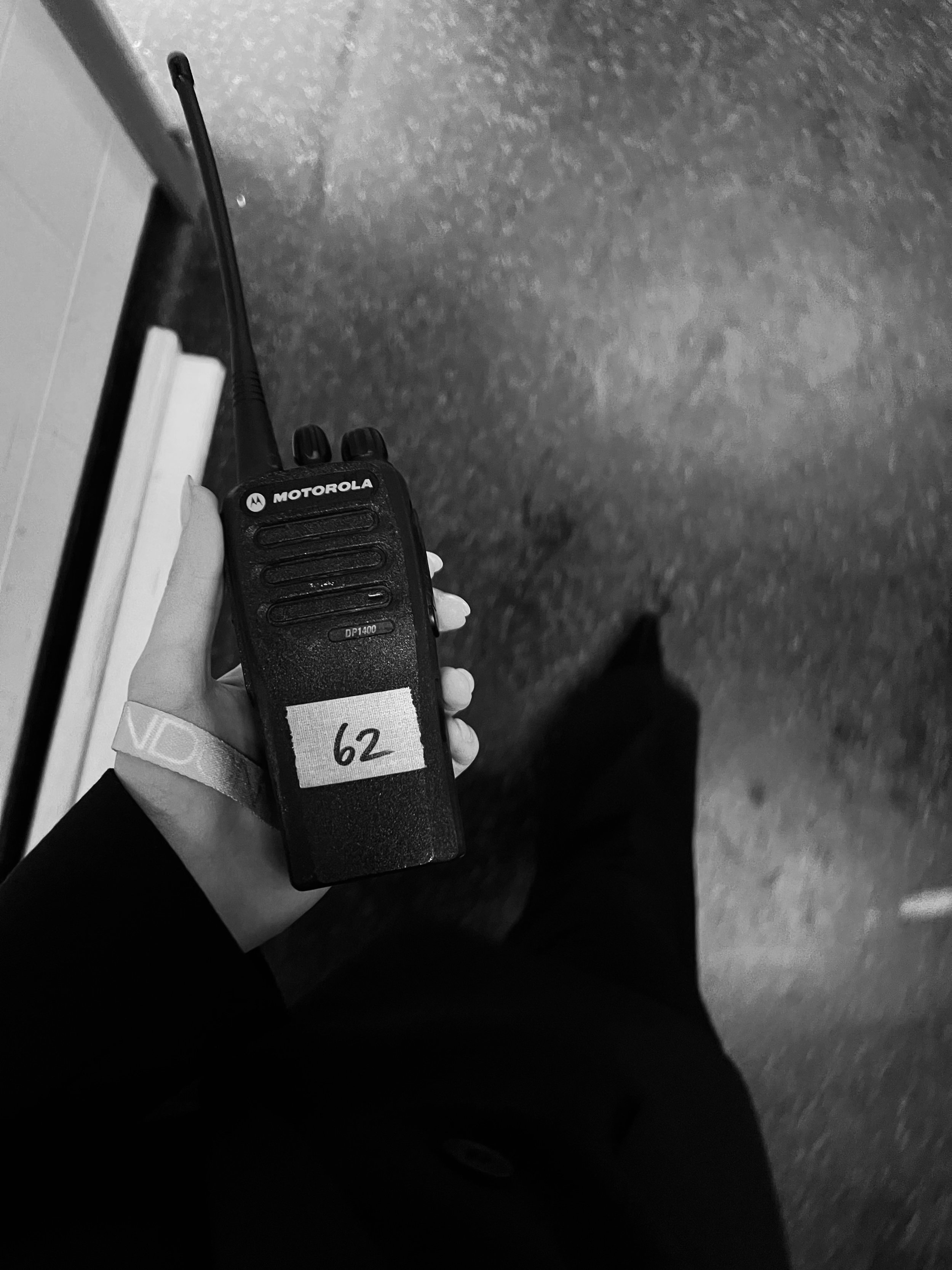 Person holding a Motorola walkie-talkie with a white label marked '62' in black text, with dark carpeted floor and a portion of a wall or door in the background.
